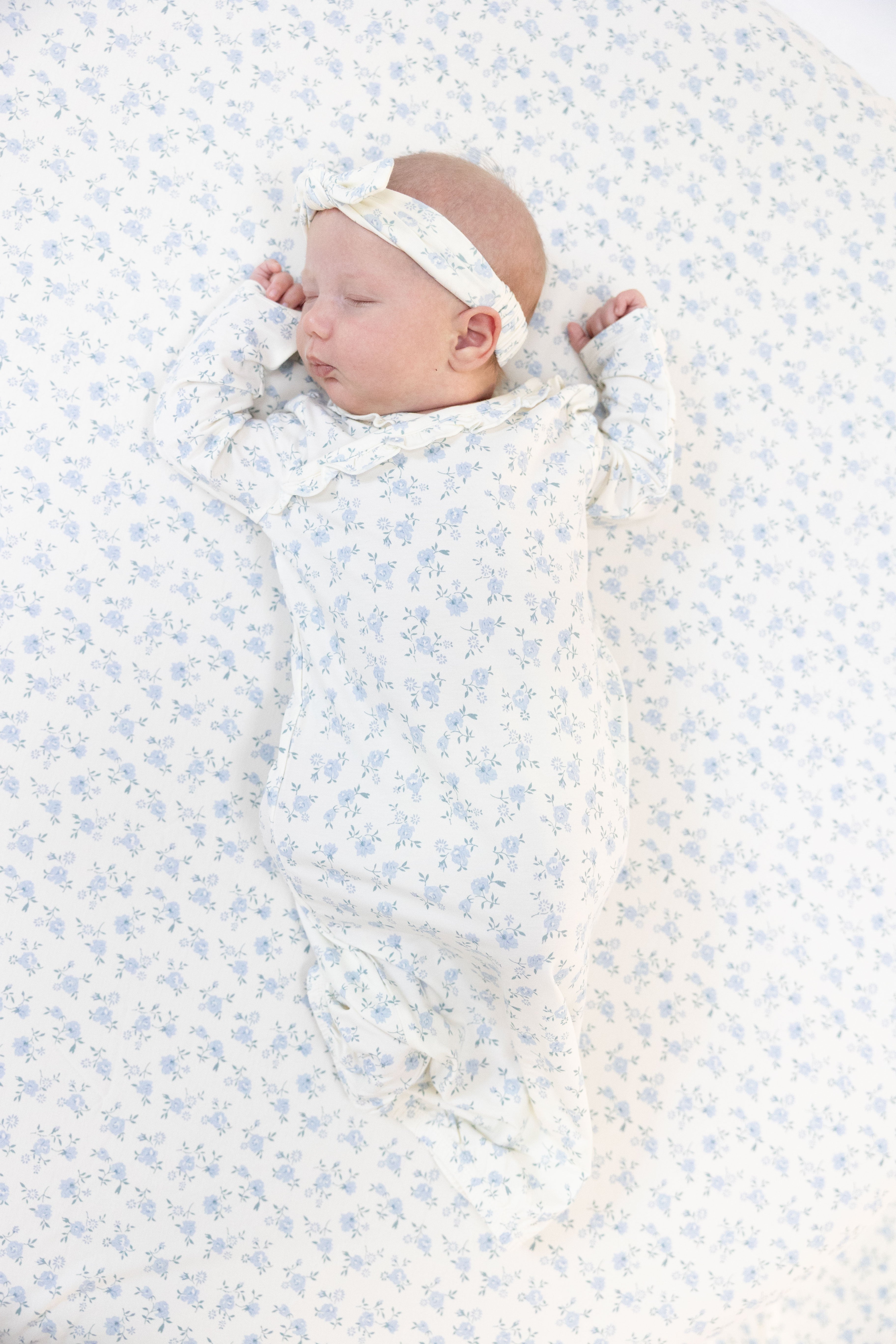 A sleeping newborn baby wearing a white long-sleeve gown and matching headband with a delicate light blue floral pattern, lying on a matching floral swaddle blanket.