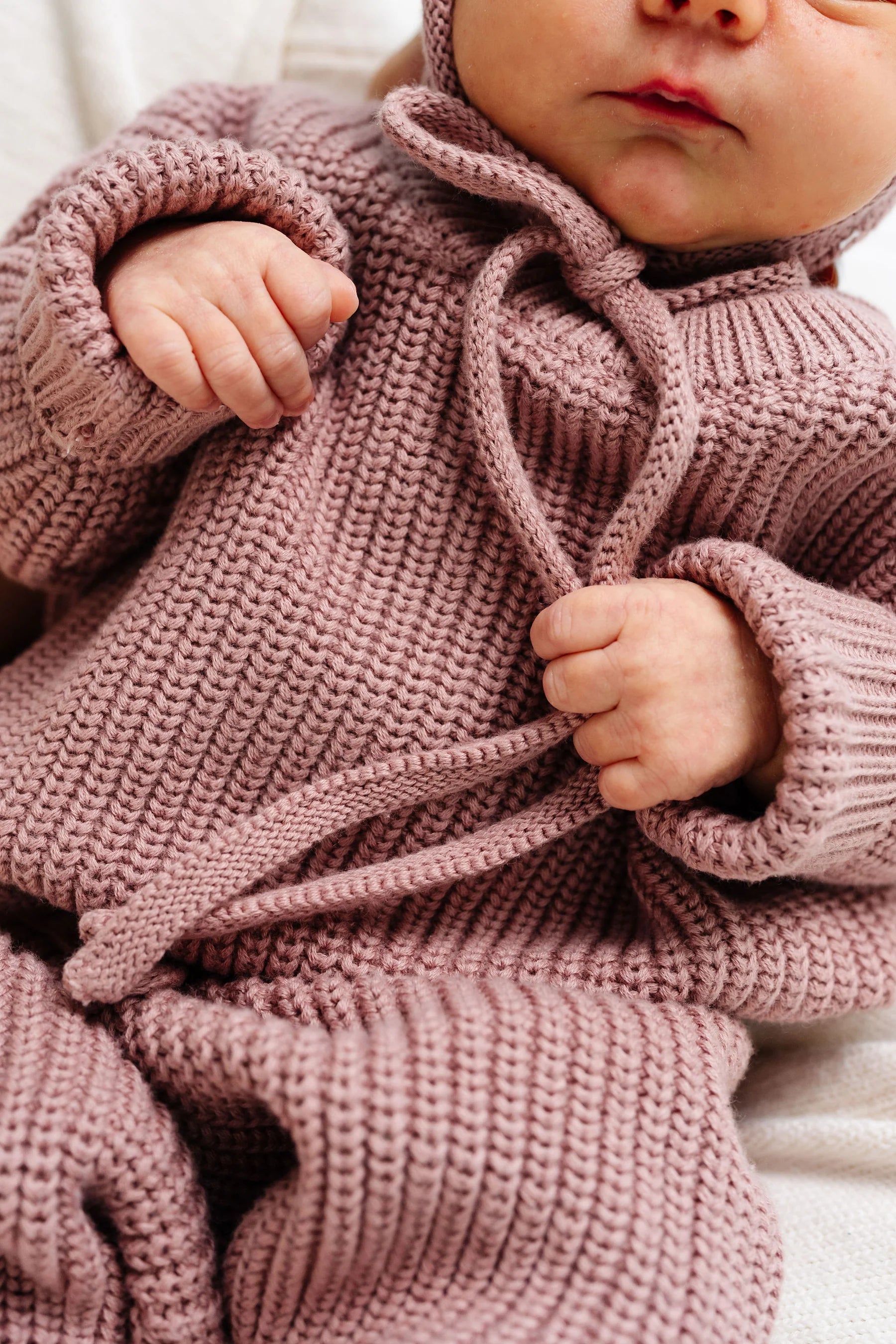 A close-up of a baby wearing a dusty rose pink chunky knit hooded romper, holding the drawstrings with both hands.