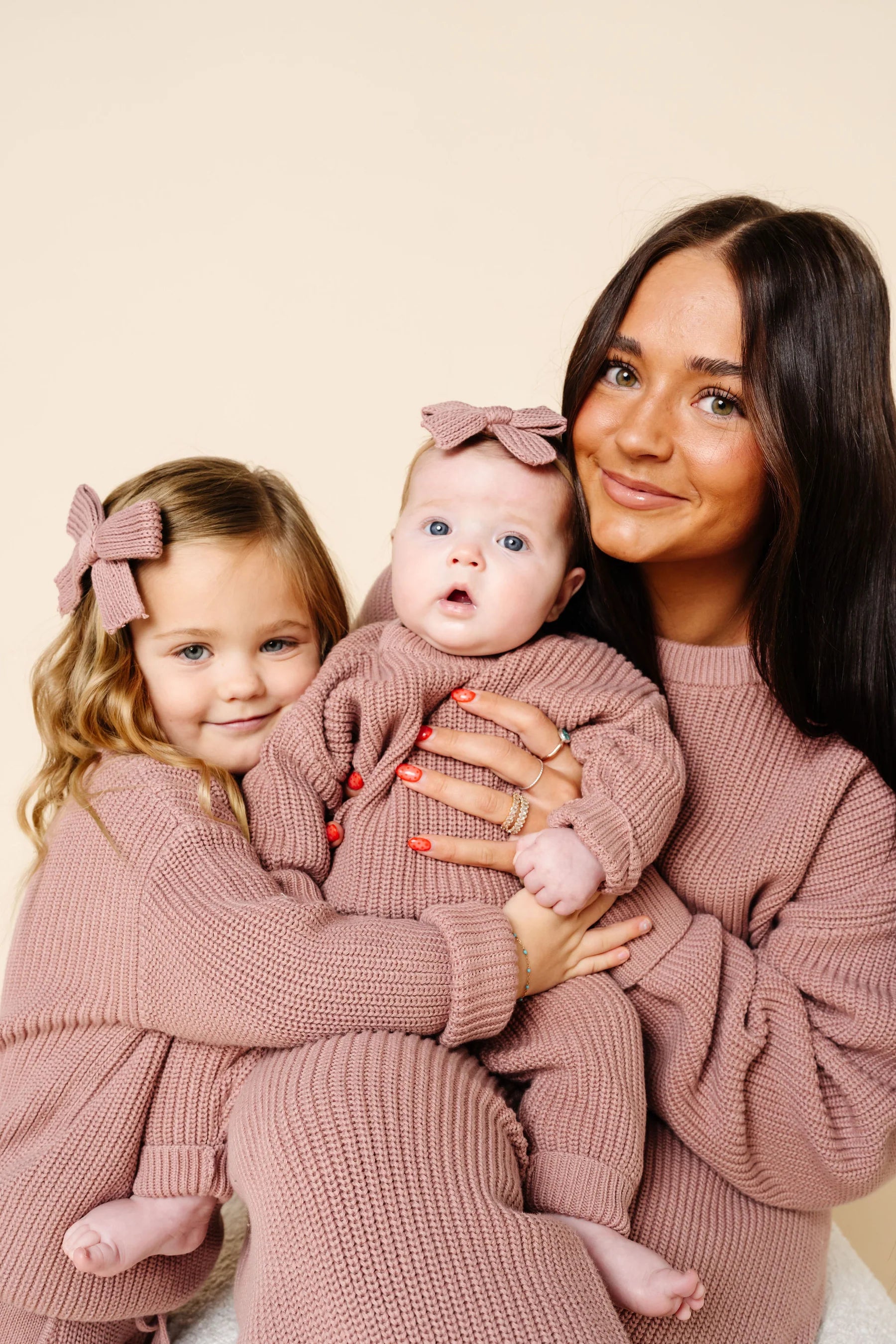 Woman holding a baby and a toddler both dressed in matching pink outfits against a beige background