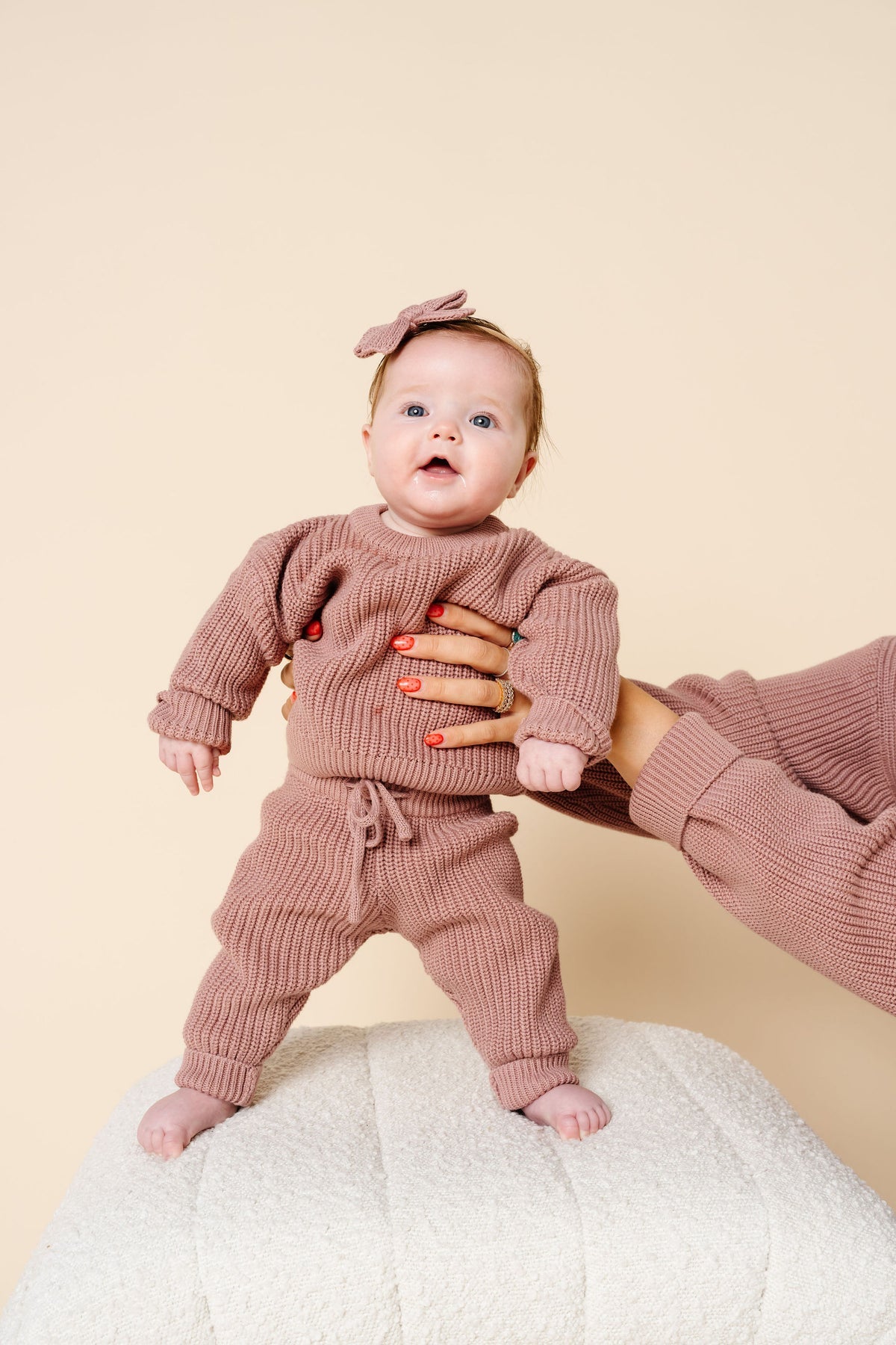A baby wearing a matching ribbed knit two-piece outfit in dusty rose pink, standing on a textured white surface while being supported by an adult's hand. The outfit includes a long-sleeve top and pants with a drawstring, and the baby is wearing a matching bow headband.