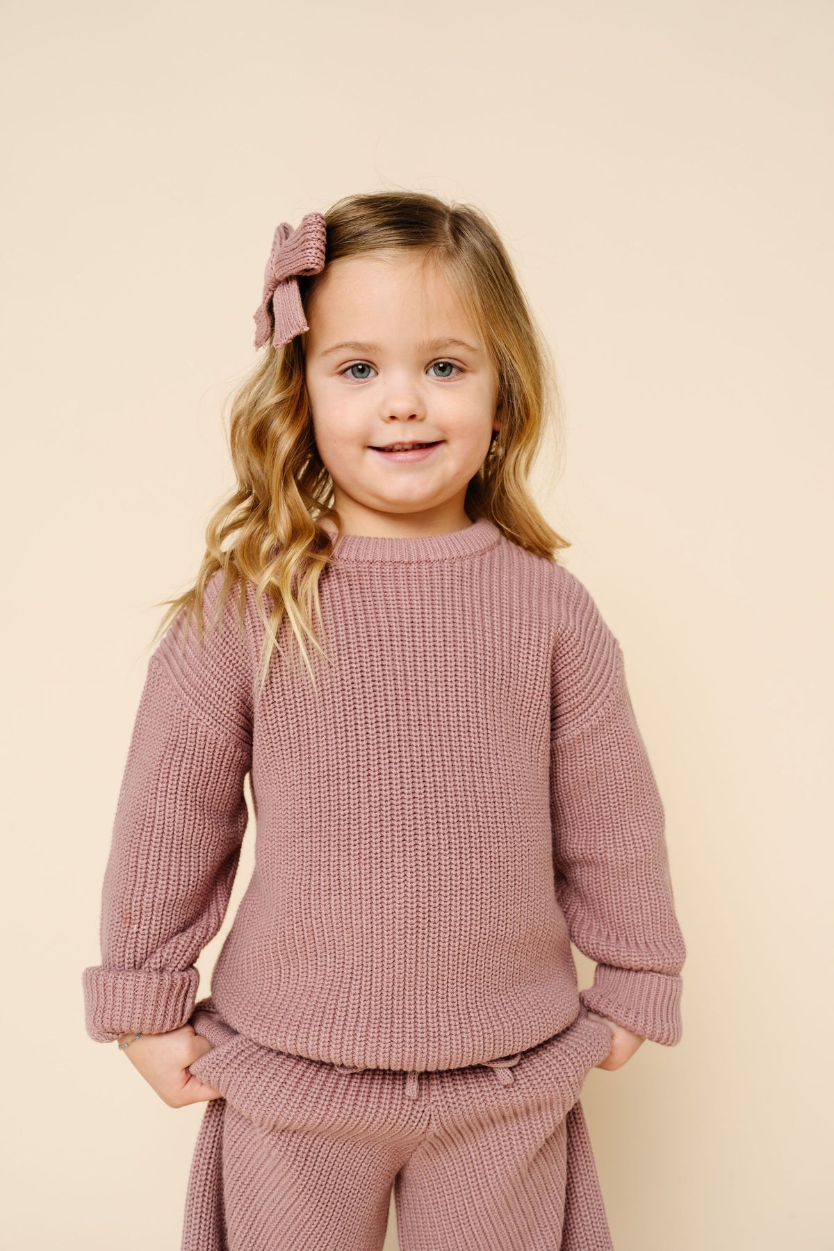 A young girl with blonde hair wearing a dusty rose ribbed knit sweater and matching pants, standing against a solid beige background.