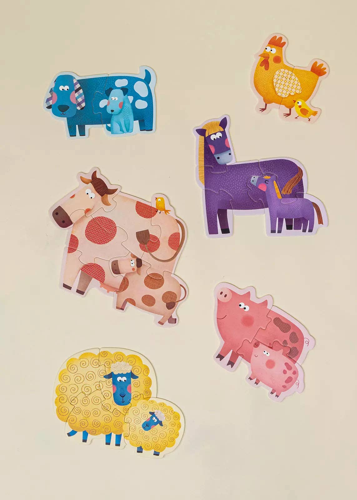 A set of colorful farm animal jigsaw puzzles, including a blue cow, yellow chicken with chick, purple horse with foal, spotted pink cow with calf, yellow sheep with blue cow, and pink pig with piglet, arranged on a light beige background.