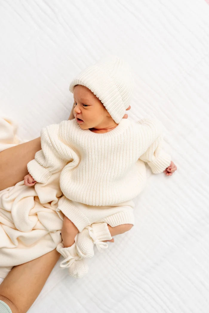 A newborn baby wearing a cream-colored ribbed knit sweater, matching beanie, booties, and a blanket, lying on a textured white blanket.