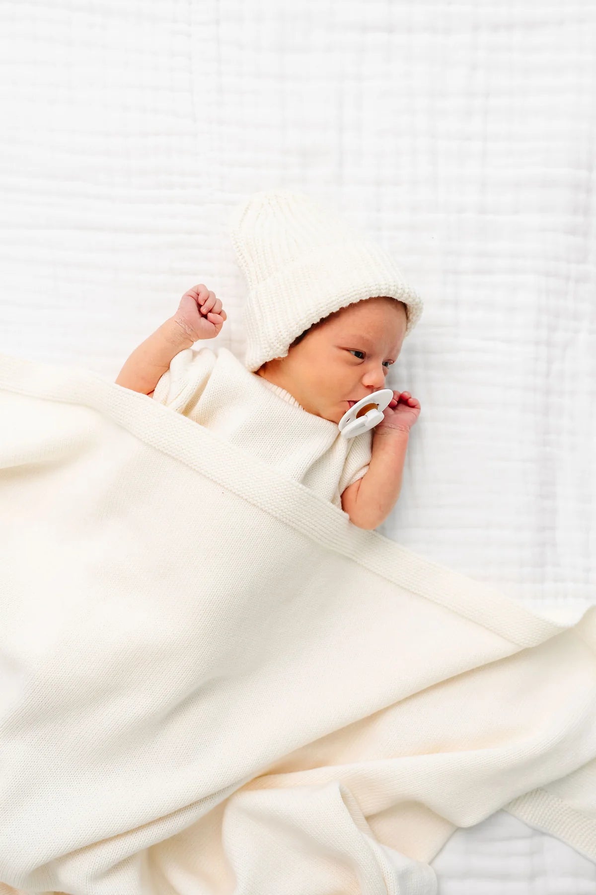 A newborn baby lying on a white textured blanket, wrapped in a soft cream blanket and wearing a matching cream knit bonnet.