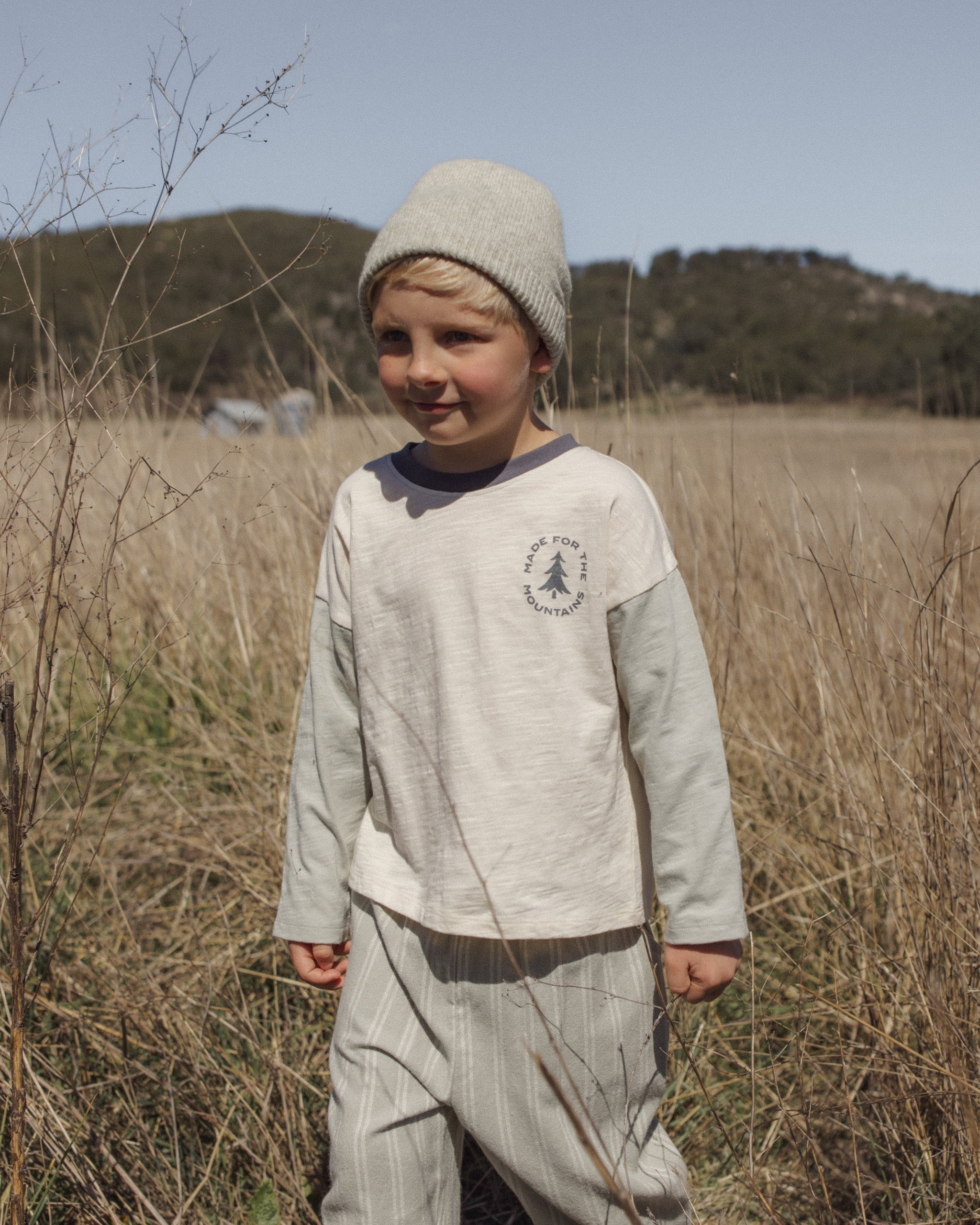 A young child standing in a dry grassy field outdoors, wearing a light beige knit beanie, a cream long-sleeve shirt with a dark chest logo, and matching light pants.