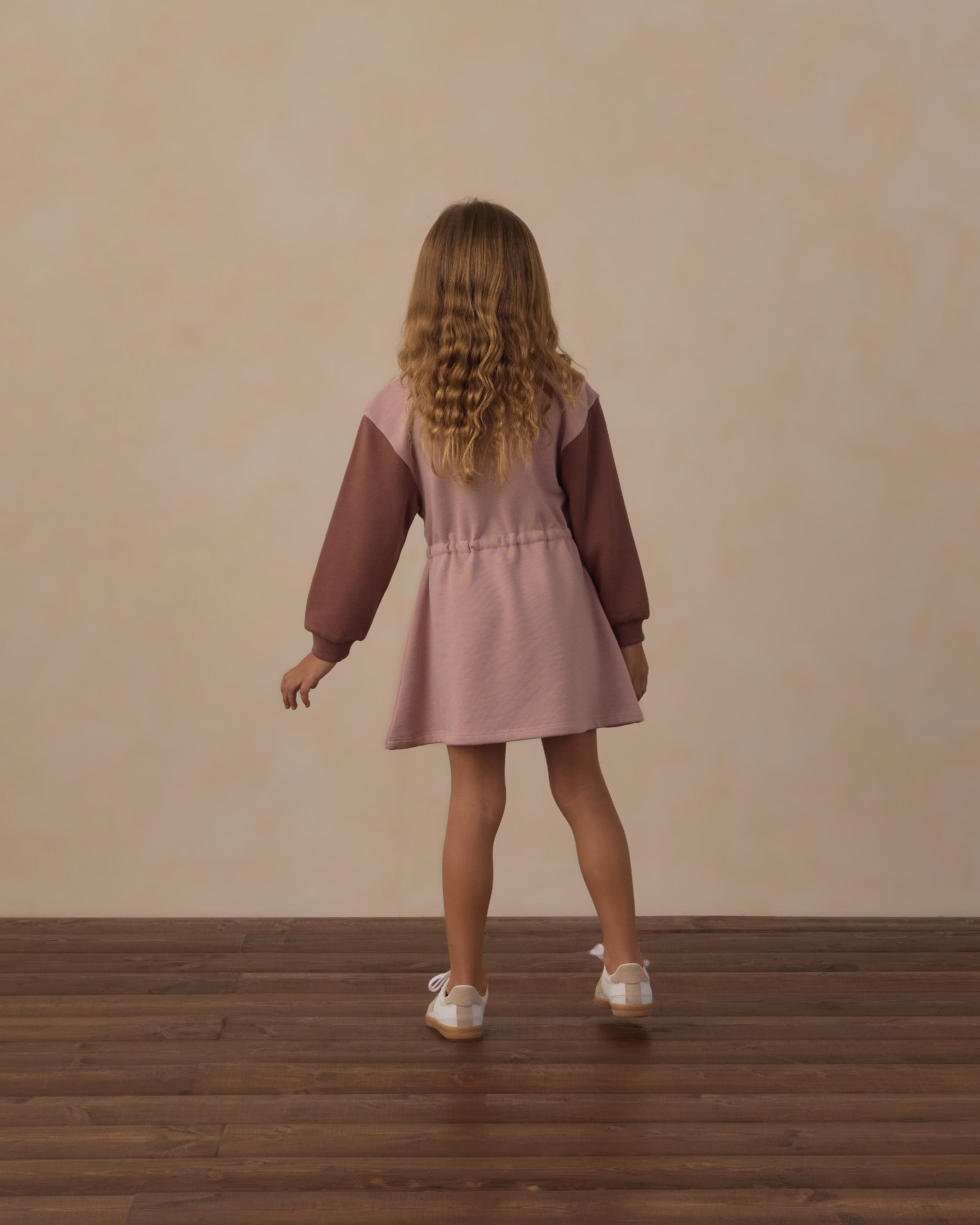 A young girl with long curly blonde hair is shown from behind, wearing a dusty pink pinafore dress with dark brown sheer long sleeves and white sneakers, standing on a dark wood floor against a textured beige wall.