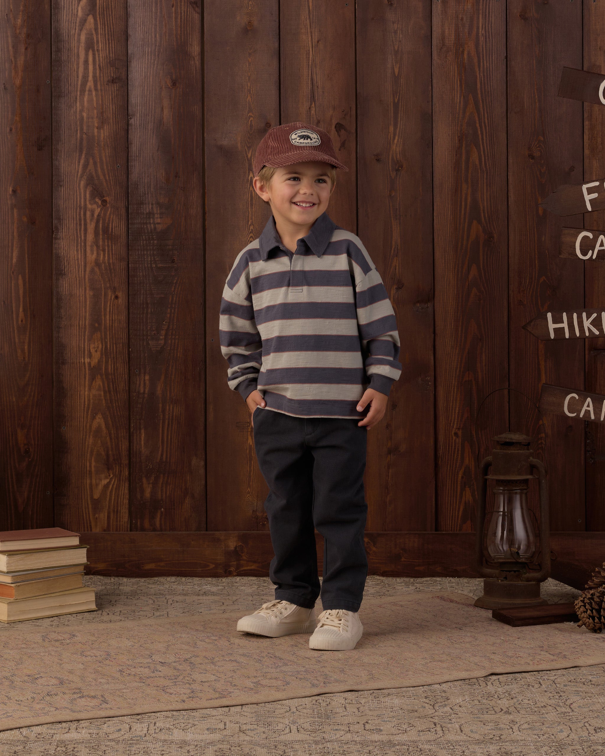 A young boy stands in front of a rustic dark wood plank wall wearing a navy and gray striped long-sleeve polo shirt, dark pants, and a brown baseball cap.