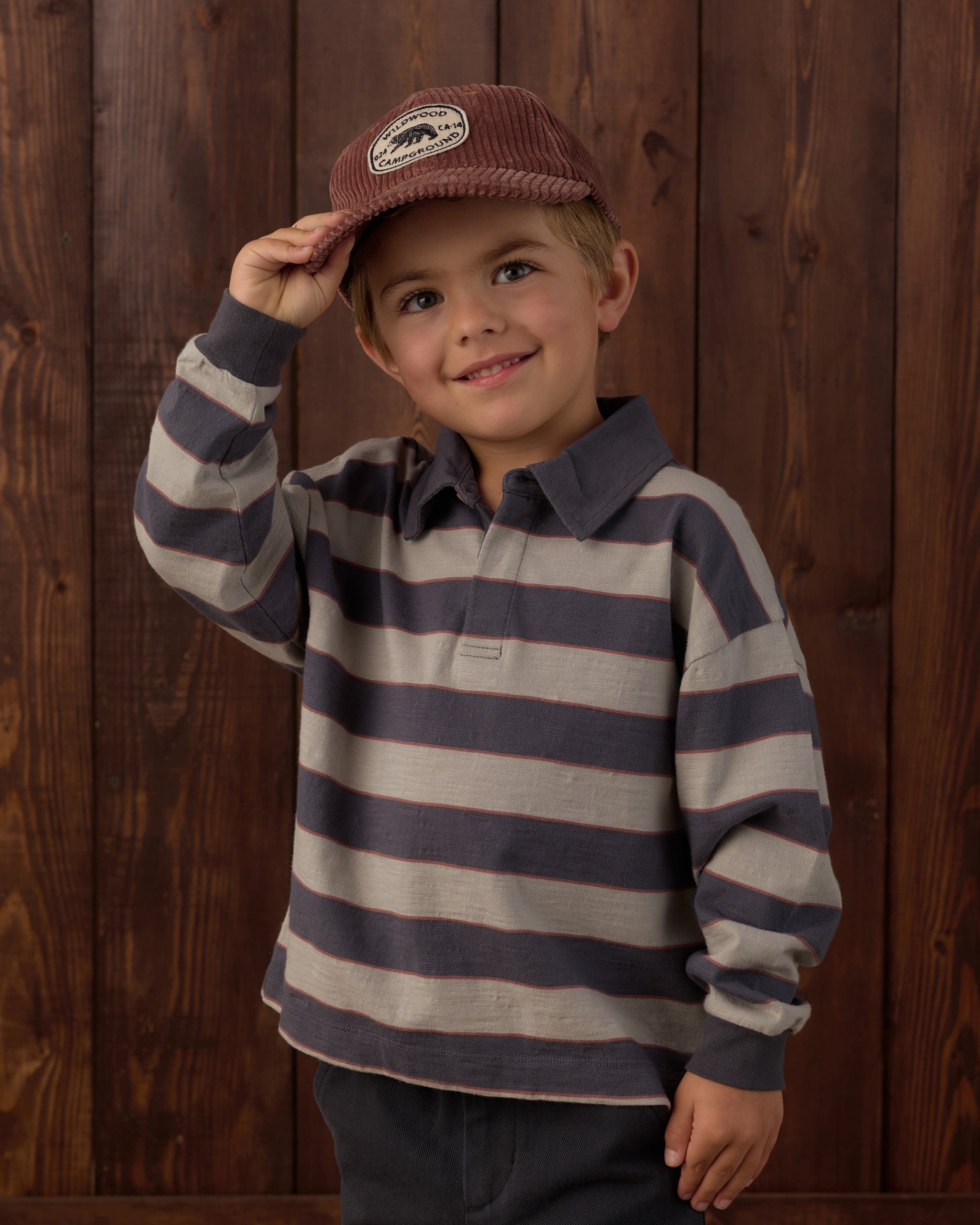 A young boy wearing a brown corduroy cap and a gray and navy striped long-sleeve polo shirt, posing in front of a dark wood plank wall.