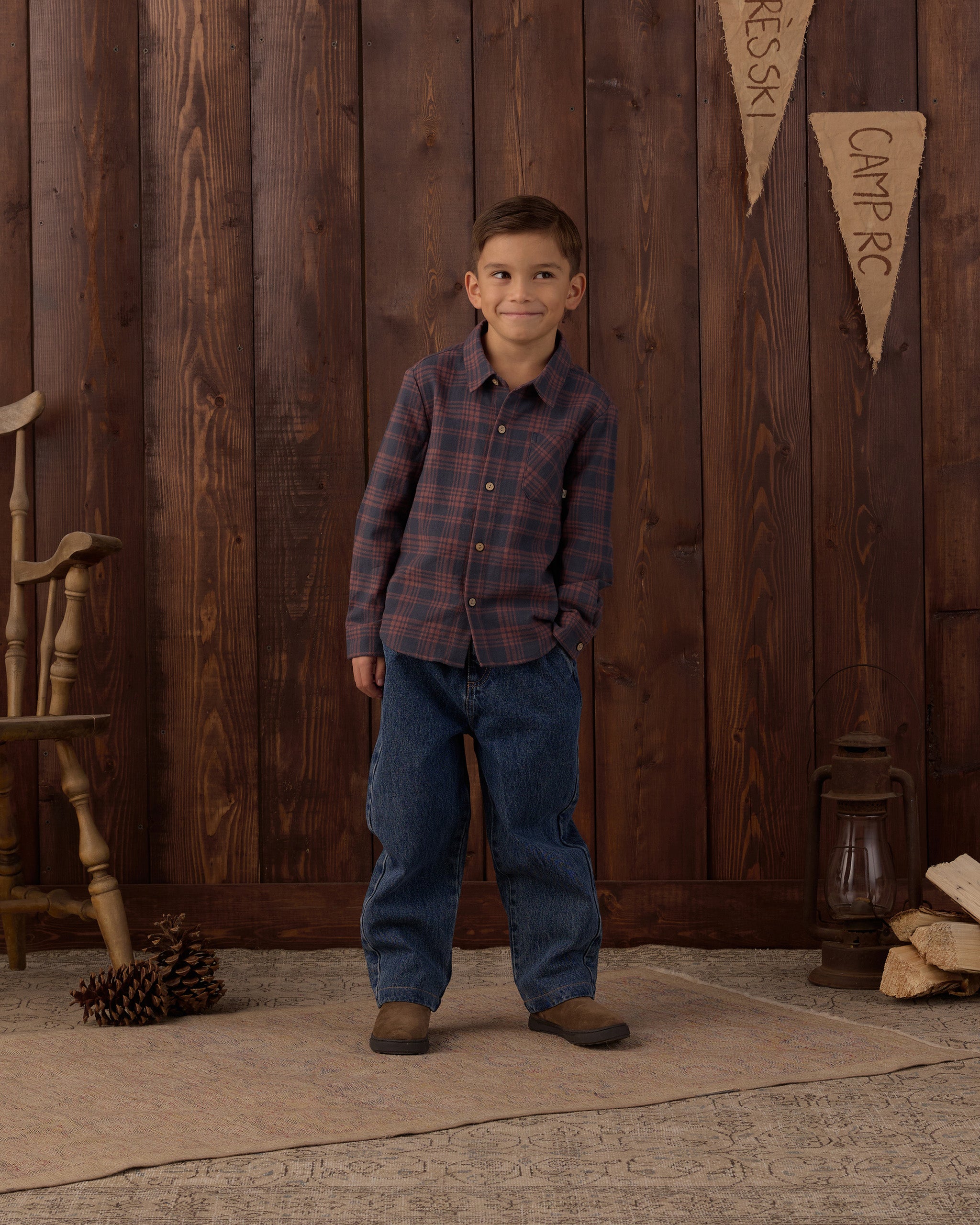 A young boy stands in front of a rustic dark wood plank wall wearing a long-sleeve plaid button-down shirt in dark red and navy, paired with blue jeans and brown boots. A wooden chair, pinecone, lantern, and stacked logs are visible in the scene.