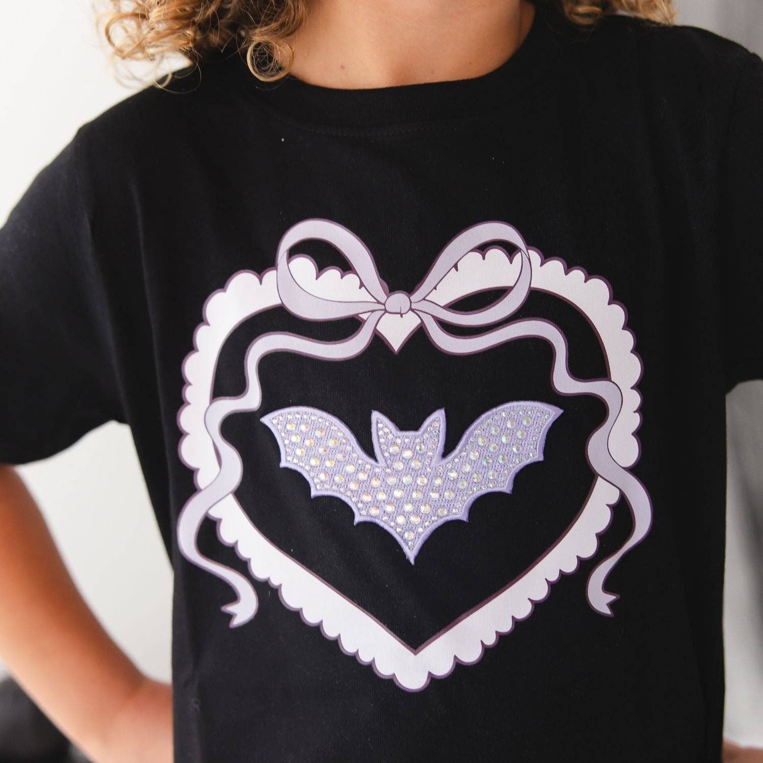 A close-up of a child wearing a black short-sleeve T-shirt with a large white heart outline, bow, and bat graphic on the front. The bat is decorated with small rhinestones.