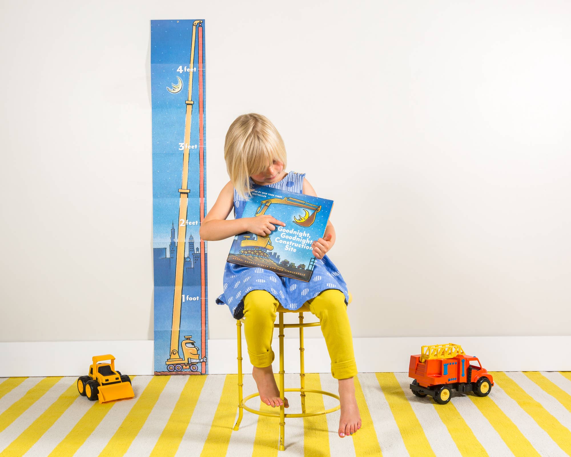 A young child sits on a yellow stool holding a book, with a tall blue growth chart featuring a construction worker and height markings standing behind them. The room has a white wall and a yellow-and-white striped rug, with toy trucks on the floor.