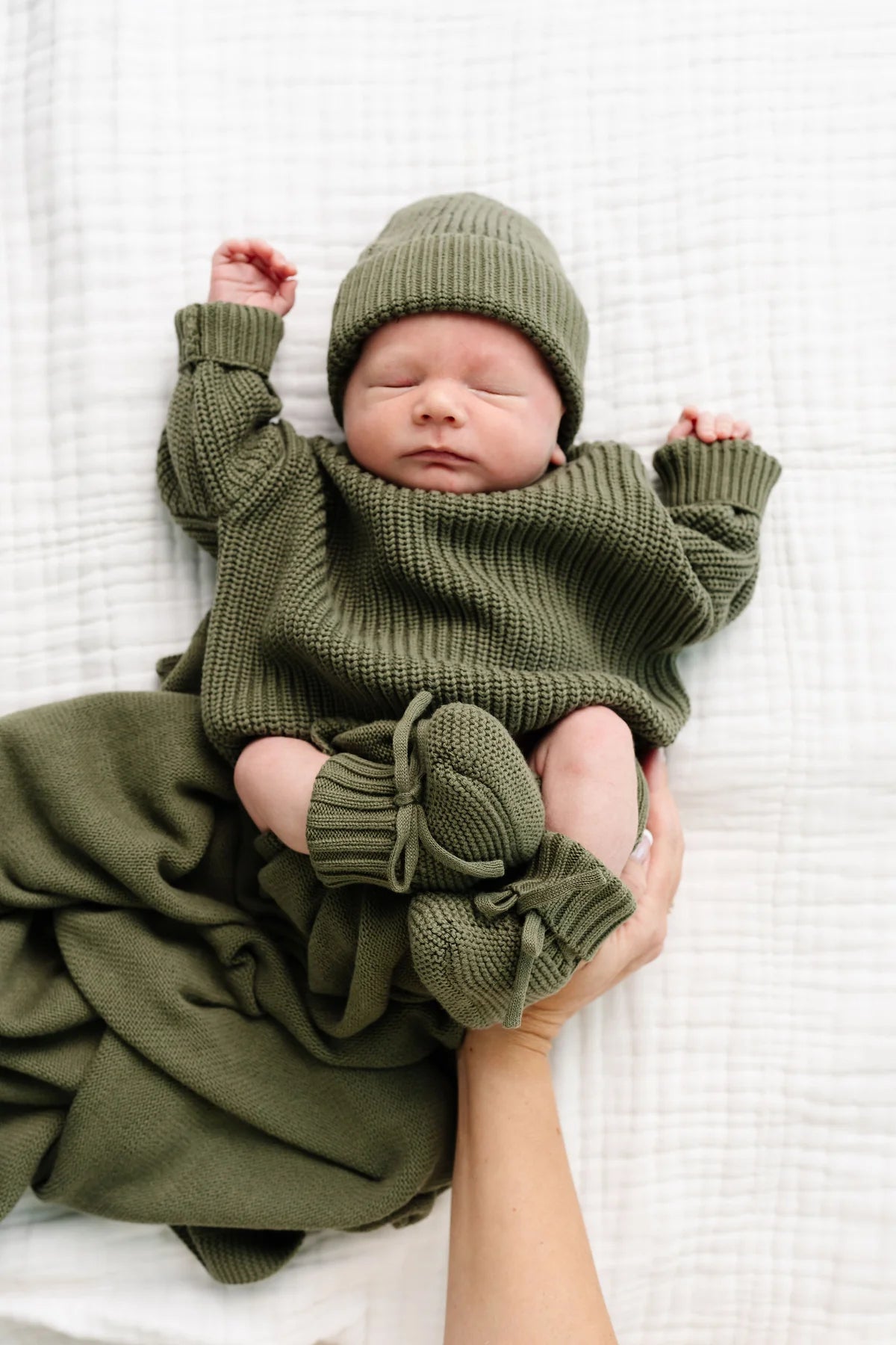 A sleeping newborn baby wearing an olive green ribbed knit sweater, matching beanie, booties, and a blanket, held gently by an adult's hand on a white textured blanket.