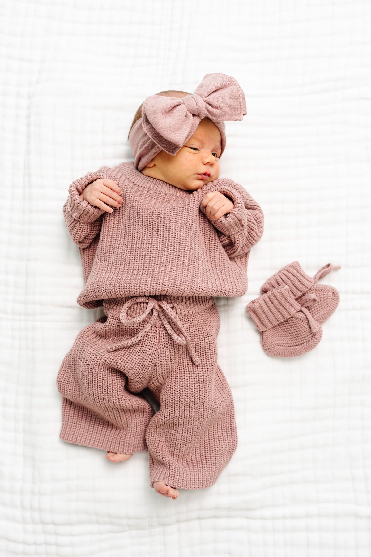 A newborn baby wearing a dusty rose knit outfit with a matching bow headband and booties, lying on a white textured blanket.