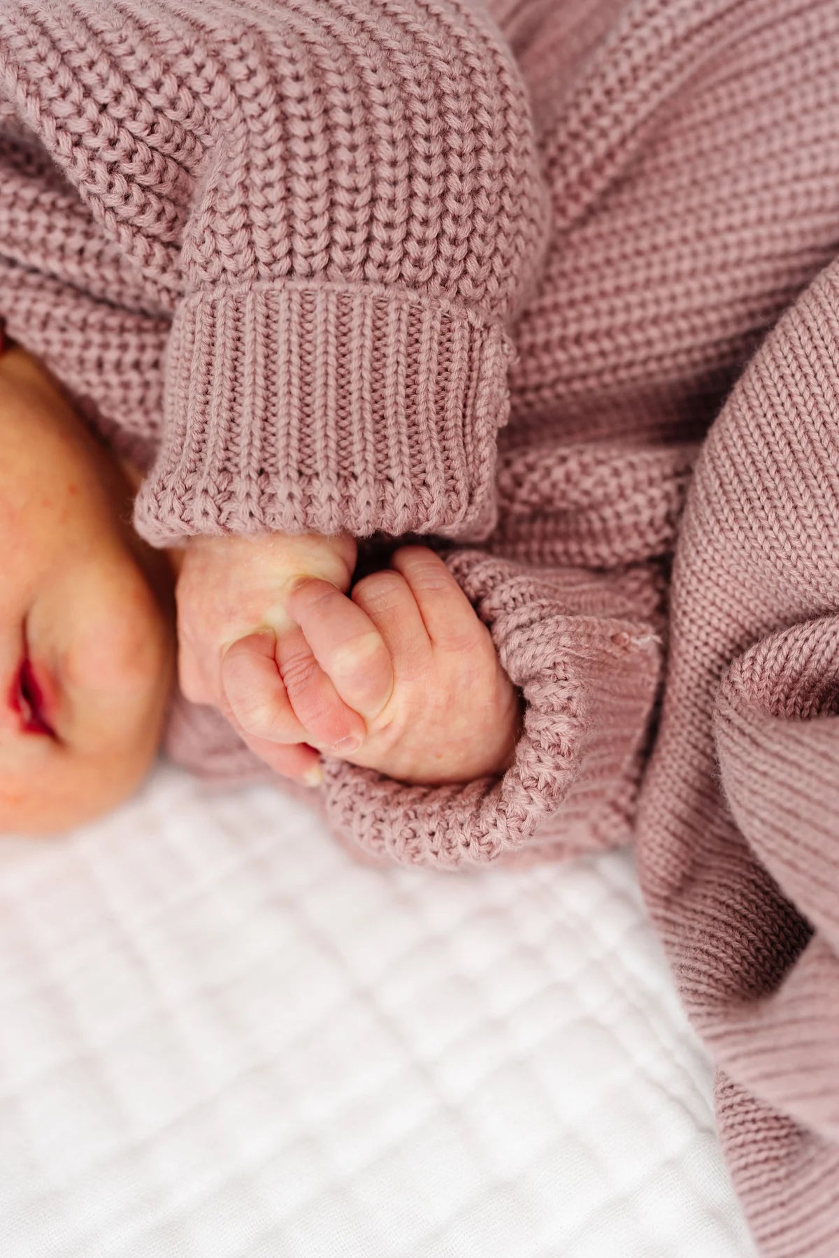 A close-up lifestyle shot of a baby wearing a dusty rose pink chunky knit blanket and matching knit clothing, holding their baby foot.
