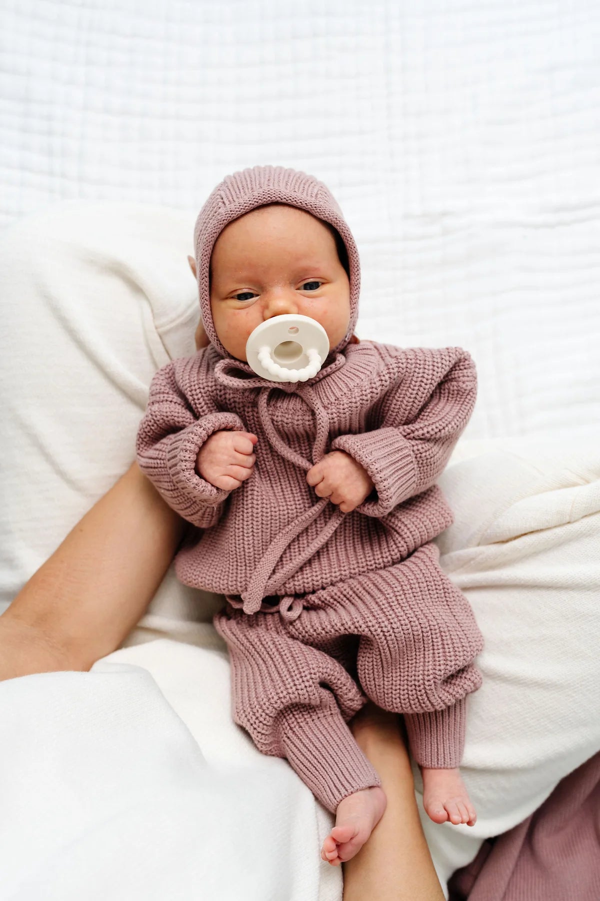 A newborn baby wearing a dusty rose pink knit hooded romper and matching pants, sitting on a person's lap while sucking a white pacifier.