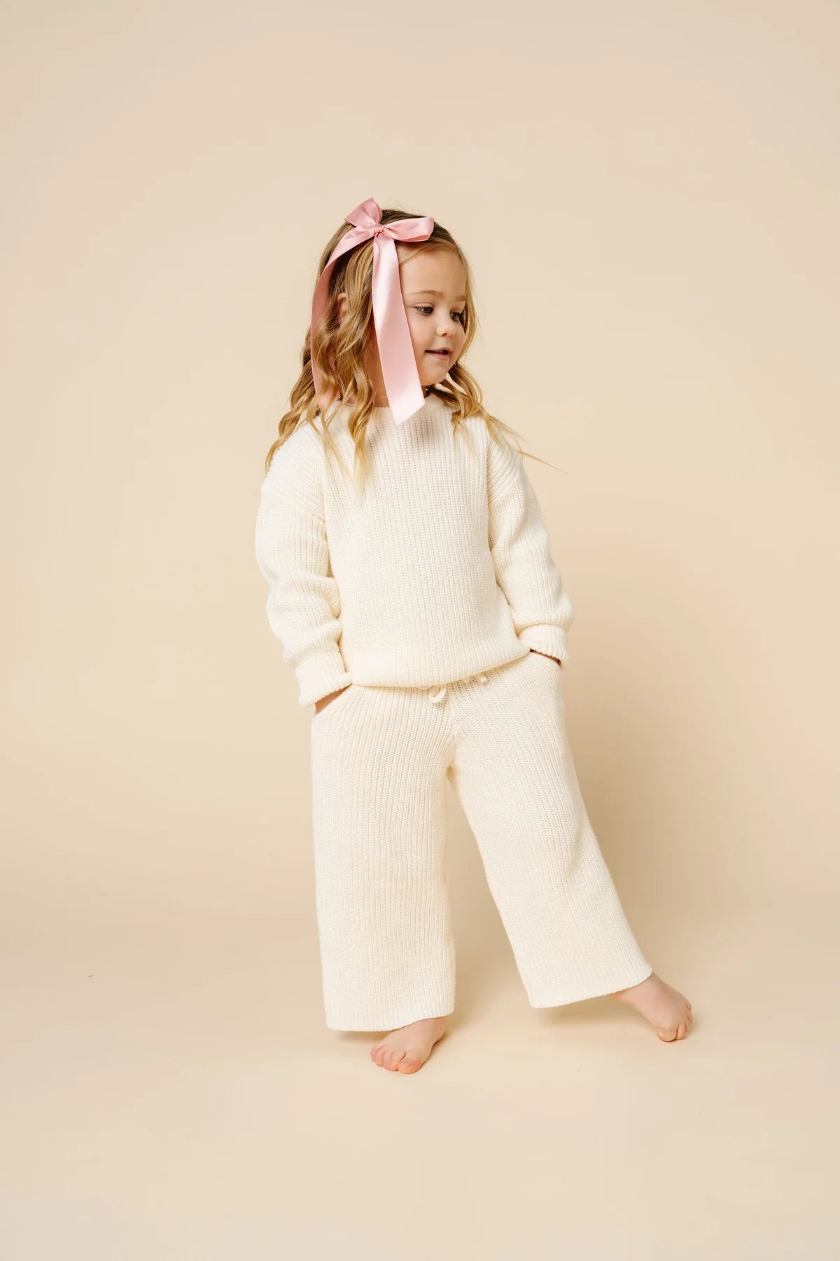 A toddler girl stands barefoot against a warm cream studio backdrop, wearing a matching cream ribbed knit sweater and wide-leg pants set with a small bow headband.