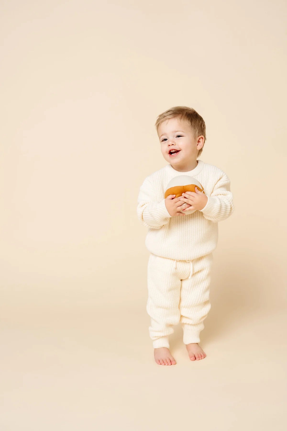 A smiling toddler standing barefoot against a warm cream studio background, wearing a matching cream knit one-piece outfit and holding a small orange toy.