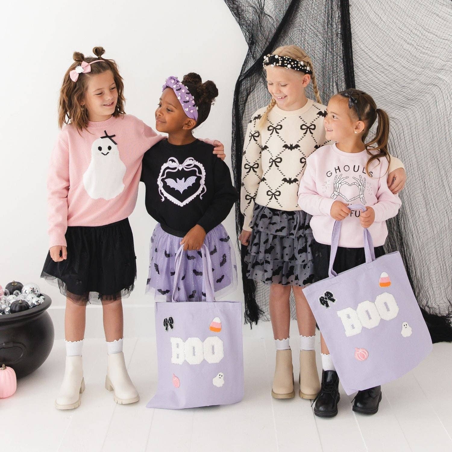 Four young girls wearing Halloween-themed sweaters and skirts stand together in a studio with black netting backdrop, each holding a lavender tote bag printed with BOO, ghosts, bats, and candy graphics.