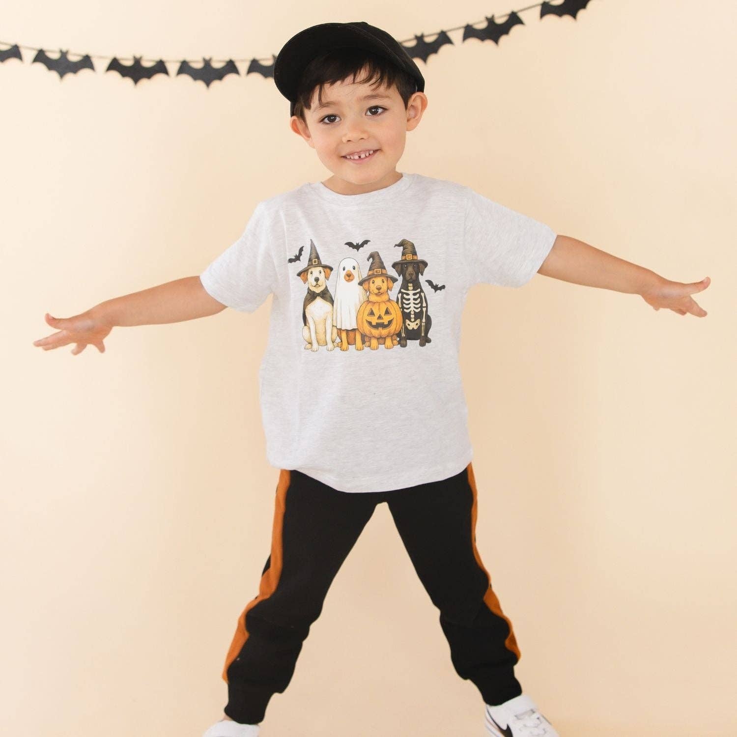 A young boy wearing a light gray heathered t-shirt with a colorful graphic of four Halloween-themed dogs, black pants with orange side stripes, and white sneakers, posing with outstretched arms against a beige background with a bat banner.