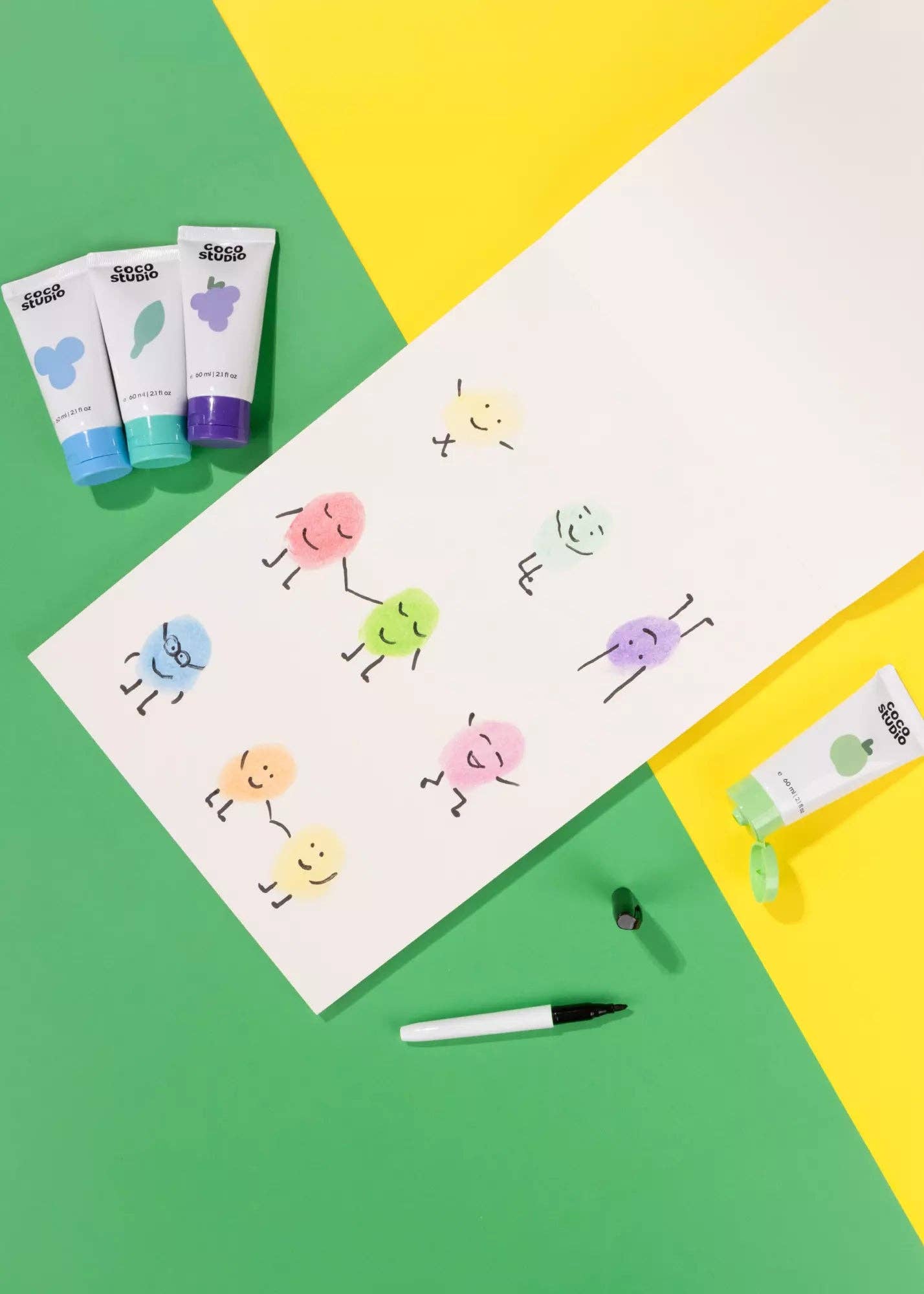 Colorful character drawings on paper with small tubes of paint and a marker on a green and yellow background.