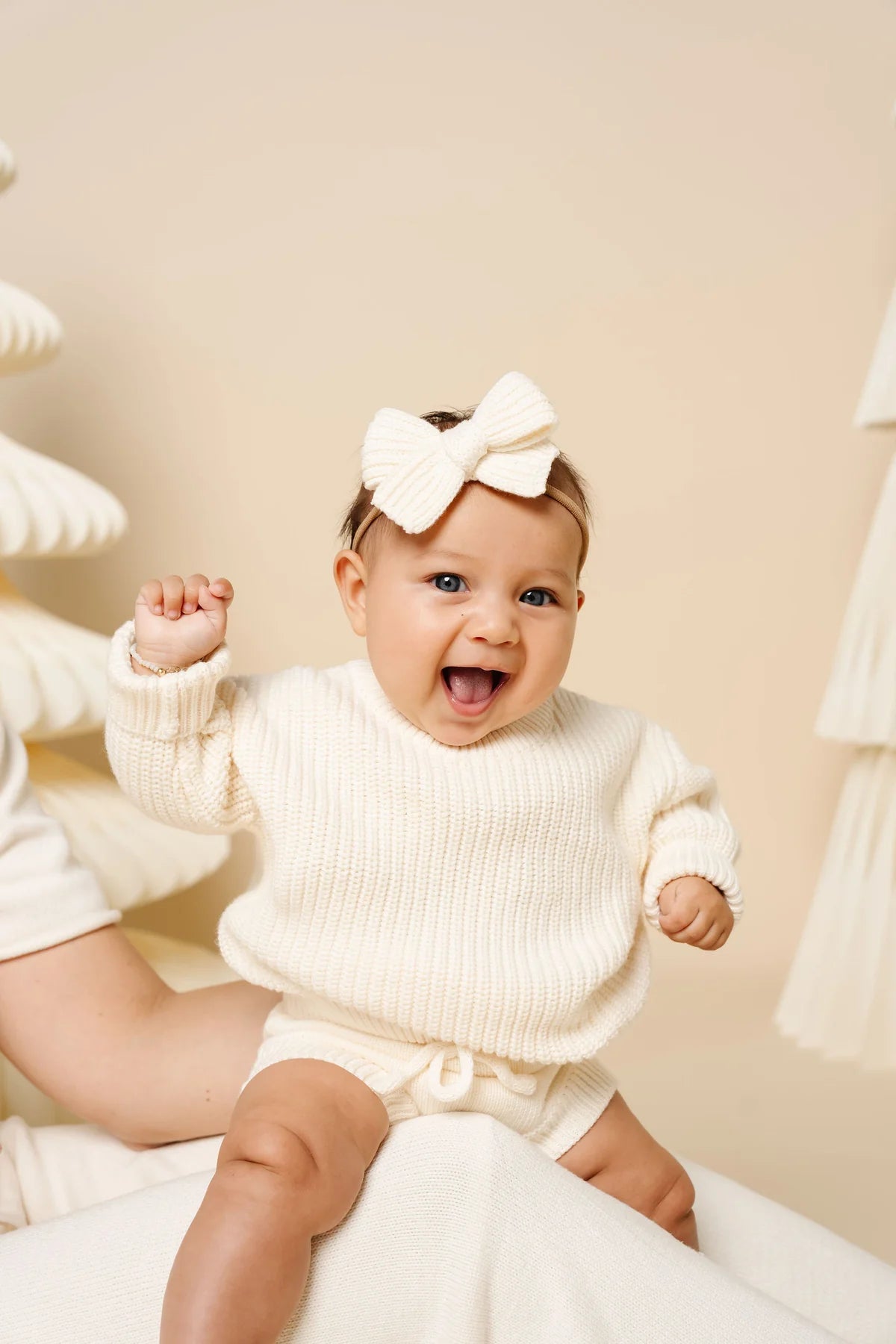 Baby wearing a white sweater and headband with a bow, sitting on a soft surface.