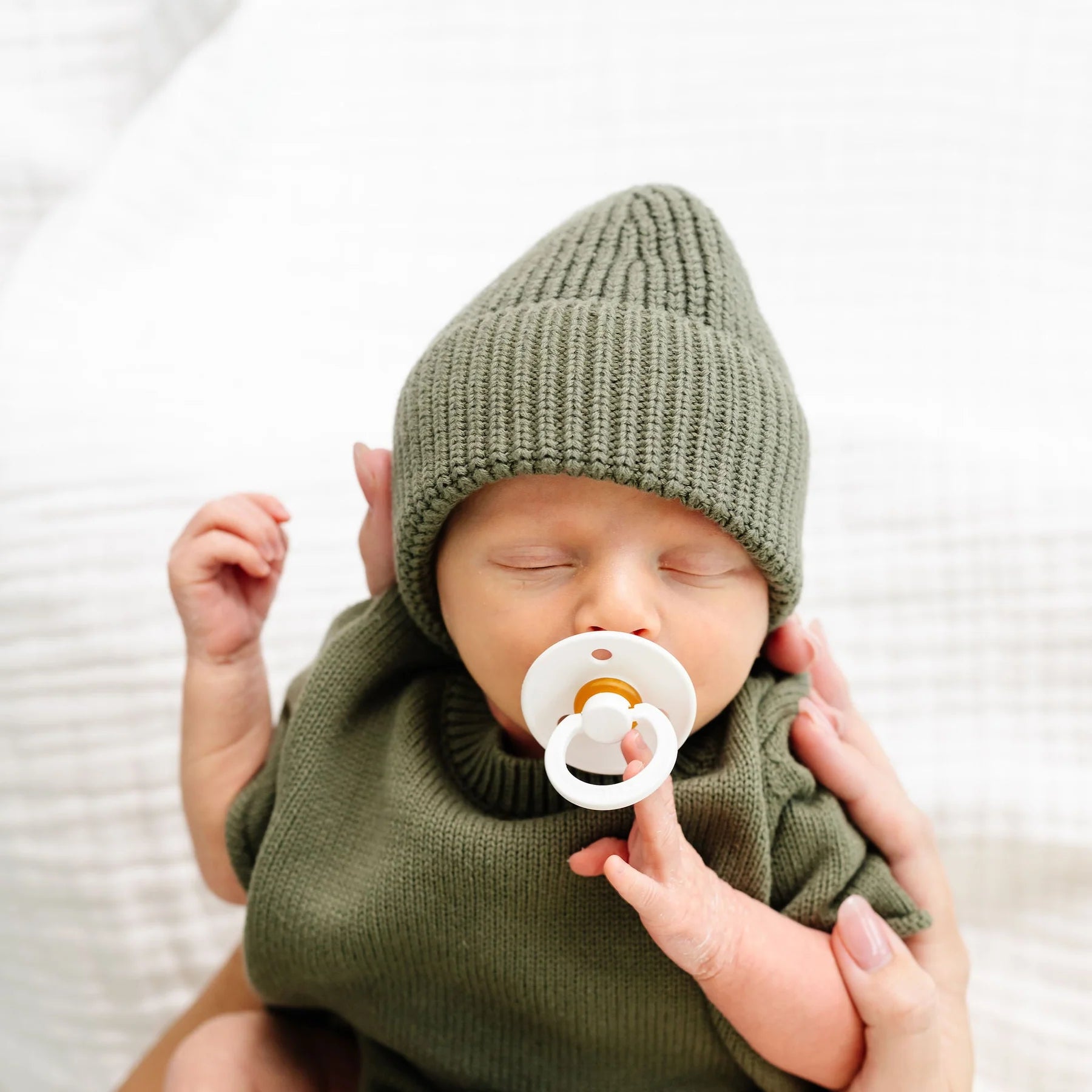 A sleeping newborn baby wearing an olive green knit bonnet and matching knit outfit, with a white pacifier in the mouth, held gently by adult hands against a soft white background.