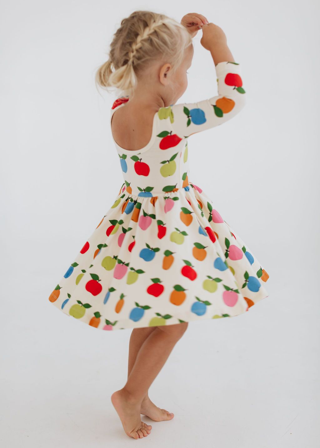 A young girl with blonde braided hair is shown from behind wearing a white long-sleeve dress with a colorful fruit print of apples, pears, and carrots, set against a plain white background.