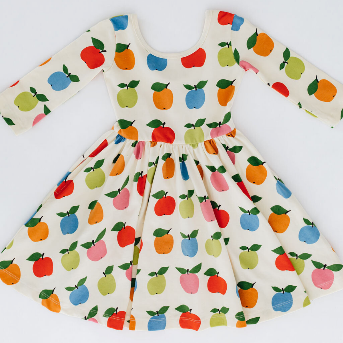 A long-sleeve baby dress with a white base and a colorful fruit print featuring red apples, orange pears, blue and green pears, and pink and red peaches. The dress has a gathered waist and a flared skirt, shown flat against a white background.