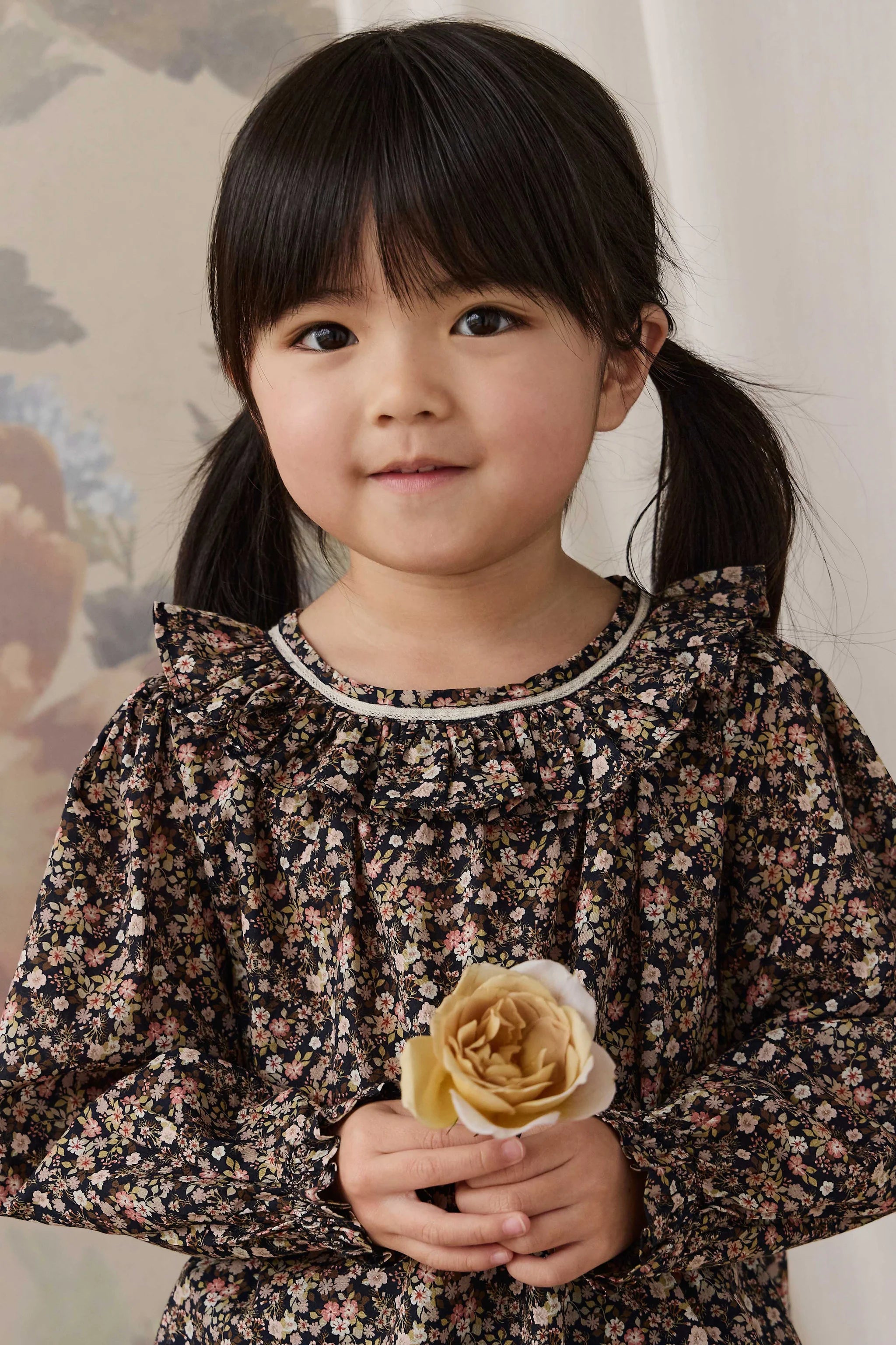 A young girl with dark hair in pigtails wearing a long-sleeve black floral dress, holding a beige rose.