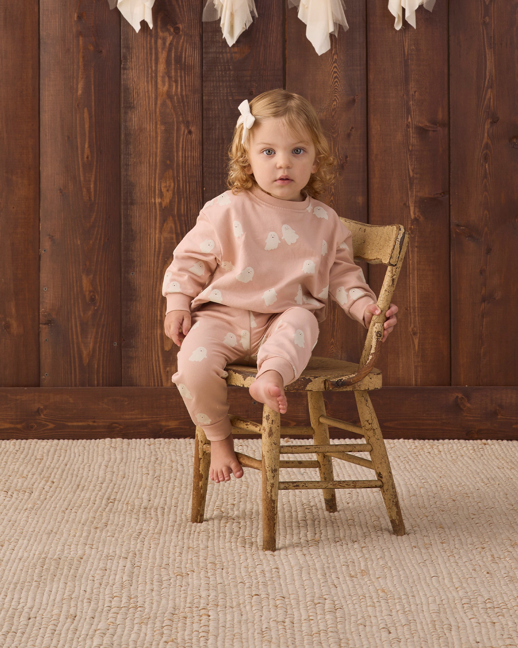 A toddler girl with blonde hair and a white bow sits on a rustic wooden chair, wearing a light pink long-sleeve pajama set with white cloud patterns. The background features a dark wood plank wall and a beige woven rug.