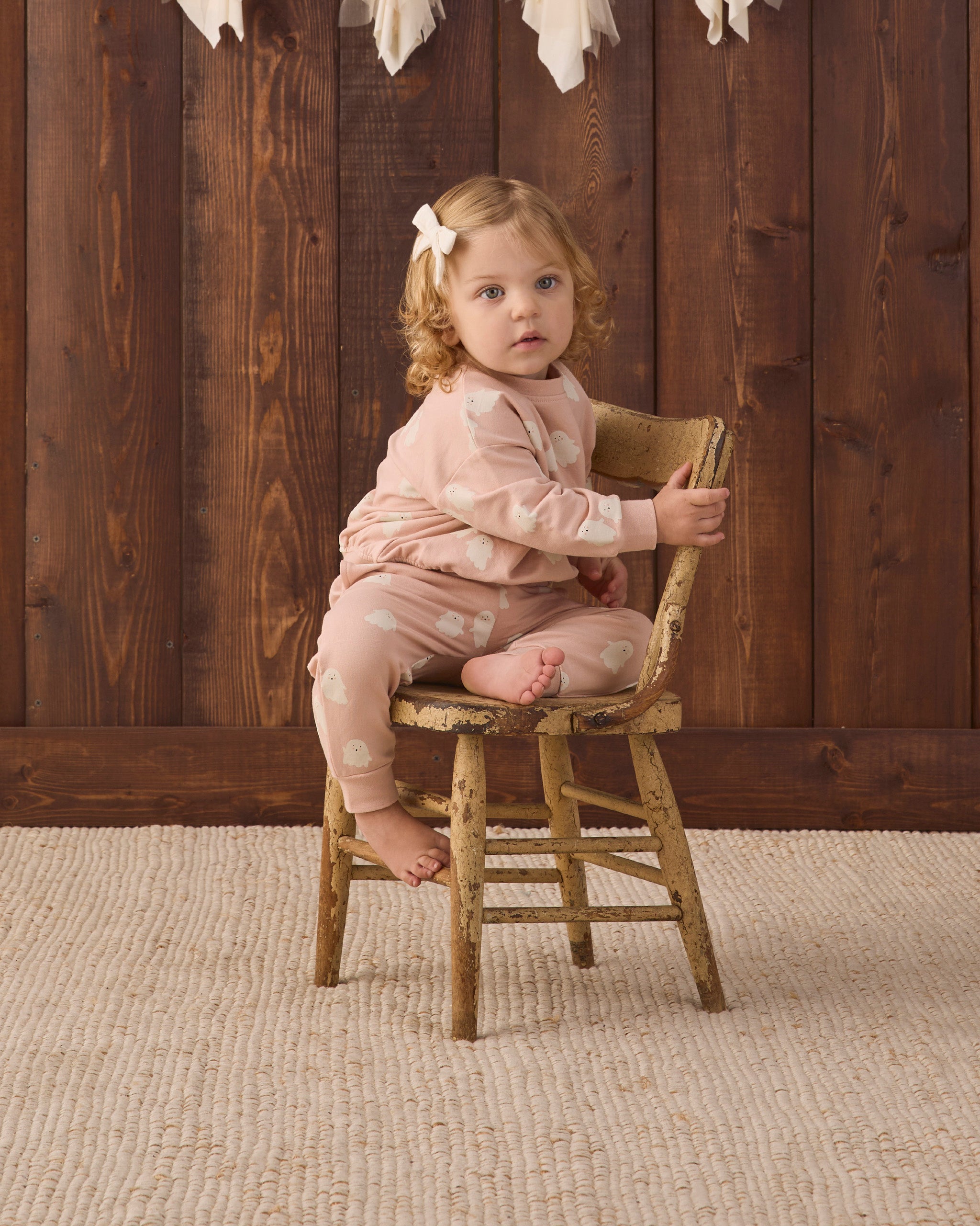 A toddler girl with curly light brown hair wearing a light pink pajama set with white polka dots sits on a rustic wooden stool in front of a dark wood plank wall.