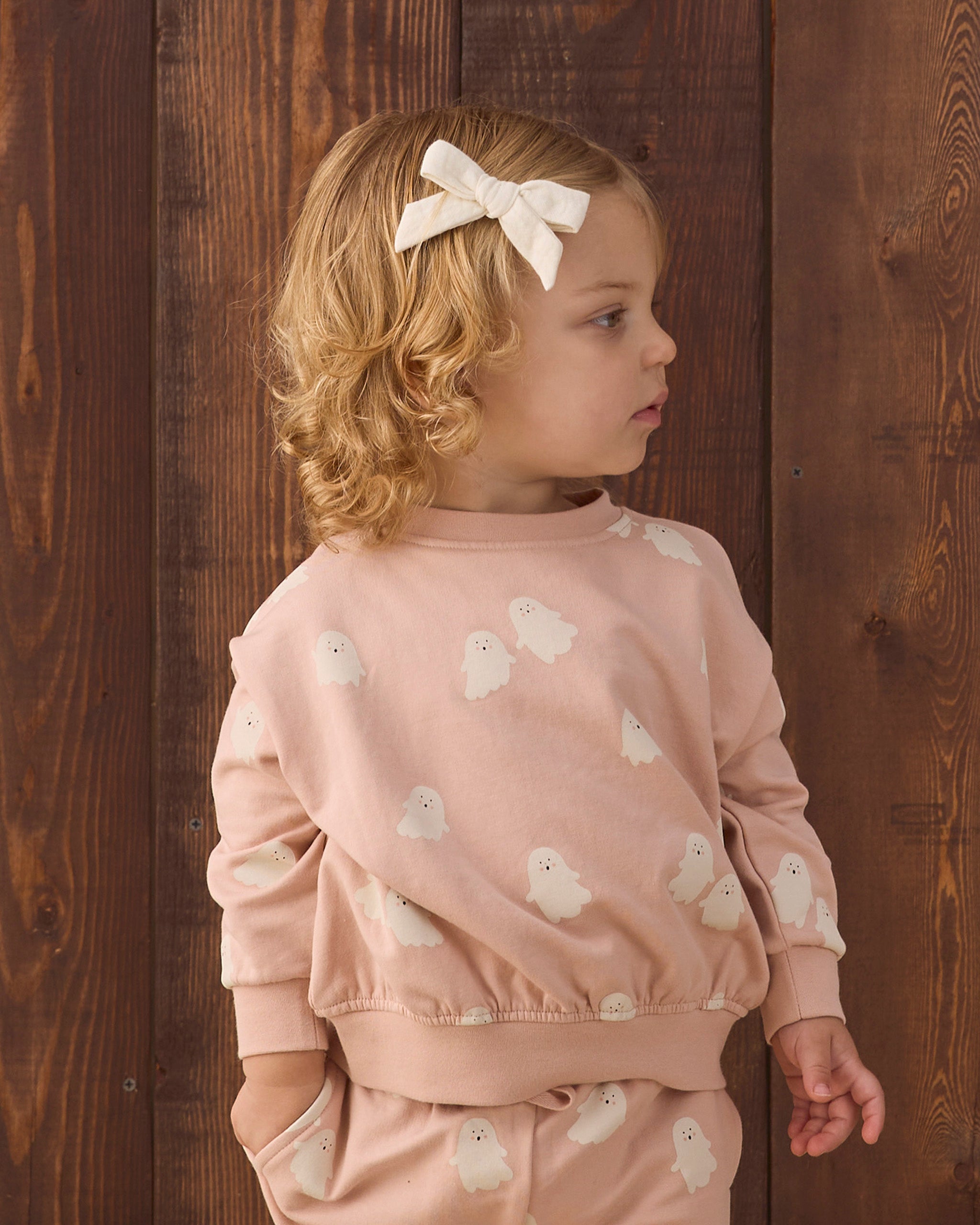 A young child with blonde curly hair wearing a light pink sweatshirt and matching pants with white ghost prints, standing against a dark wooden plank background.