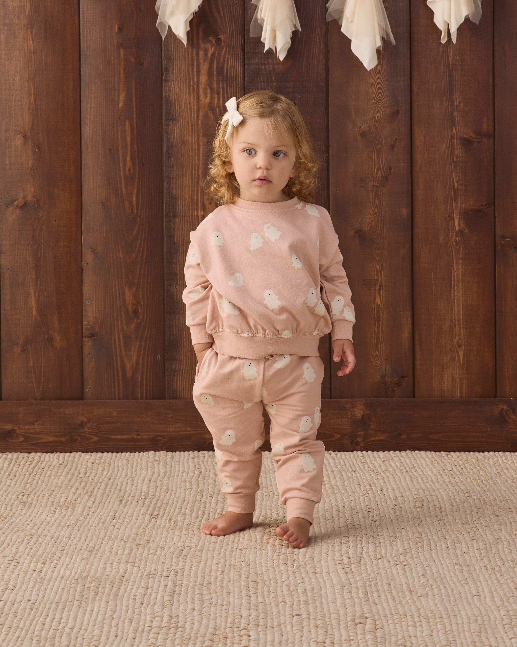 A toddler girl stands barefoot on a textured beige rug in front of a dark wood plank wall, wearing a matching dusty pink two-piece pajama set with white polka dots. The set includes a long-sleeve top and matching pants.