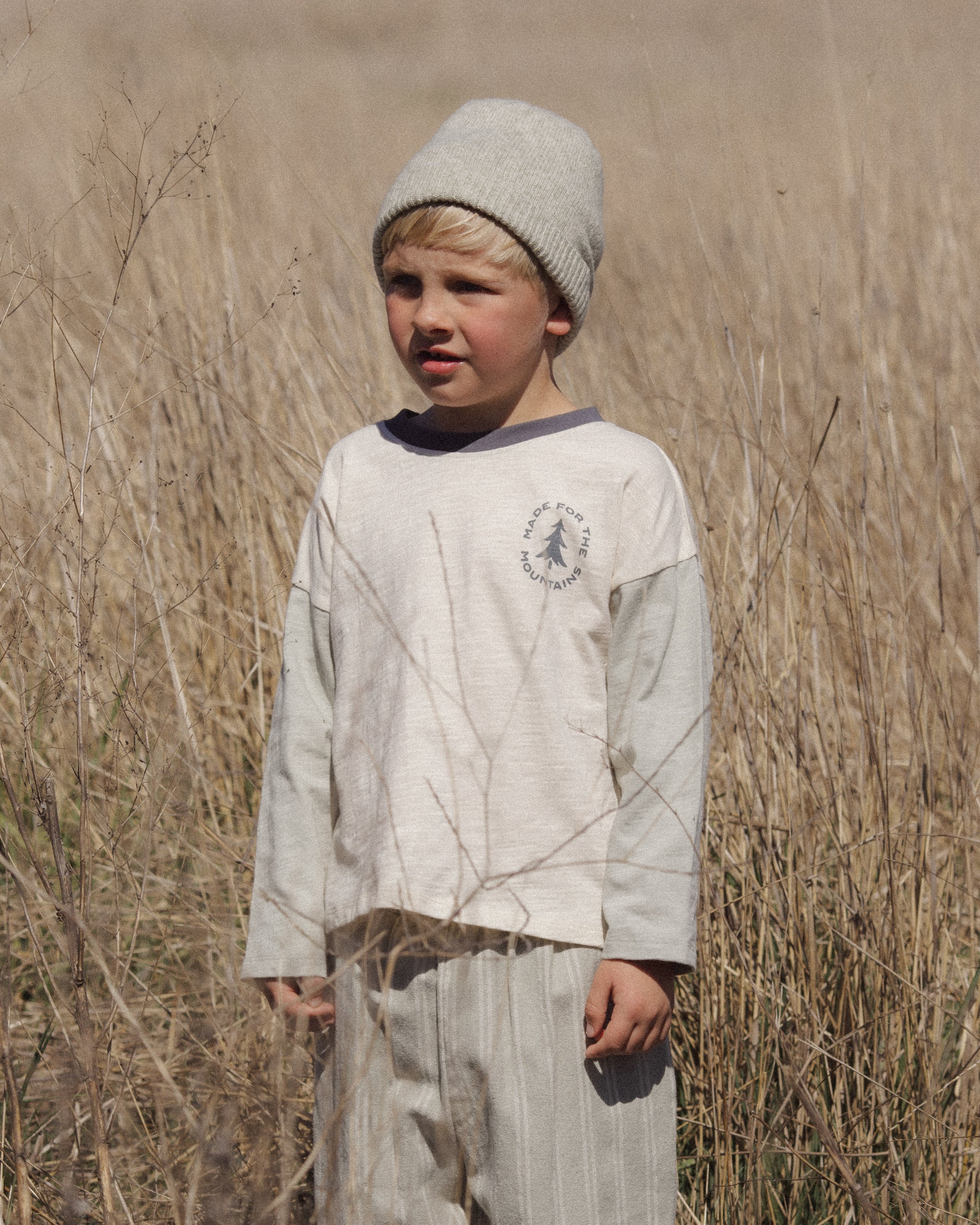 A young boy standing in a dry grass field, wearing a light gray knit beanie, a light gray long-sleeve shirt with a dark gray chest graphic, and matching light gray pants.