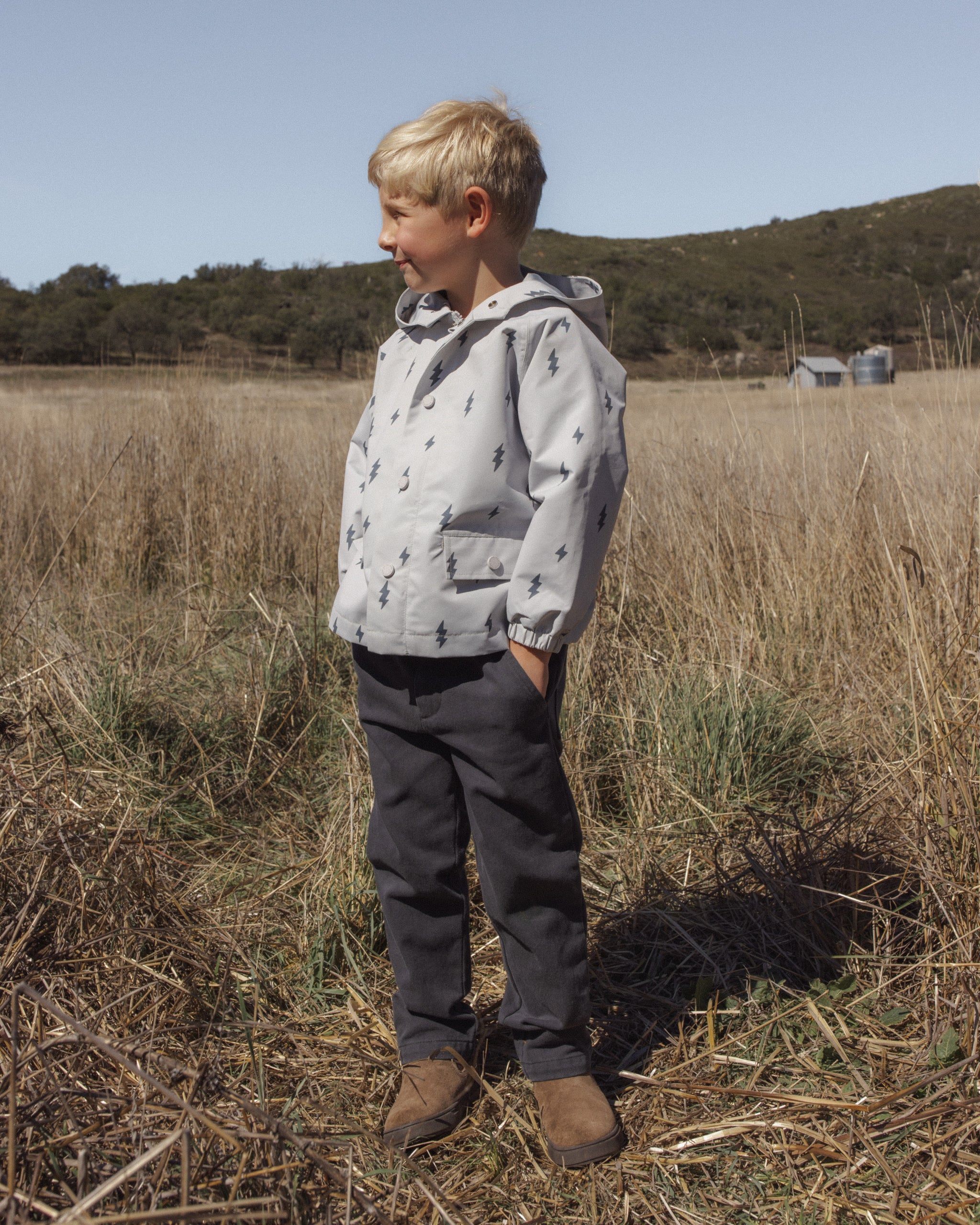 A young boy stands in a dry grassy field wearing a light gray hooded jacket with a dark tree print, dark gray pants, and brown boots.