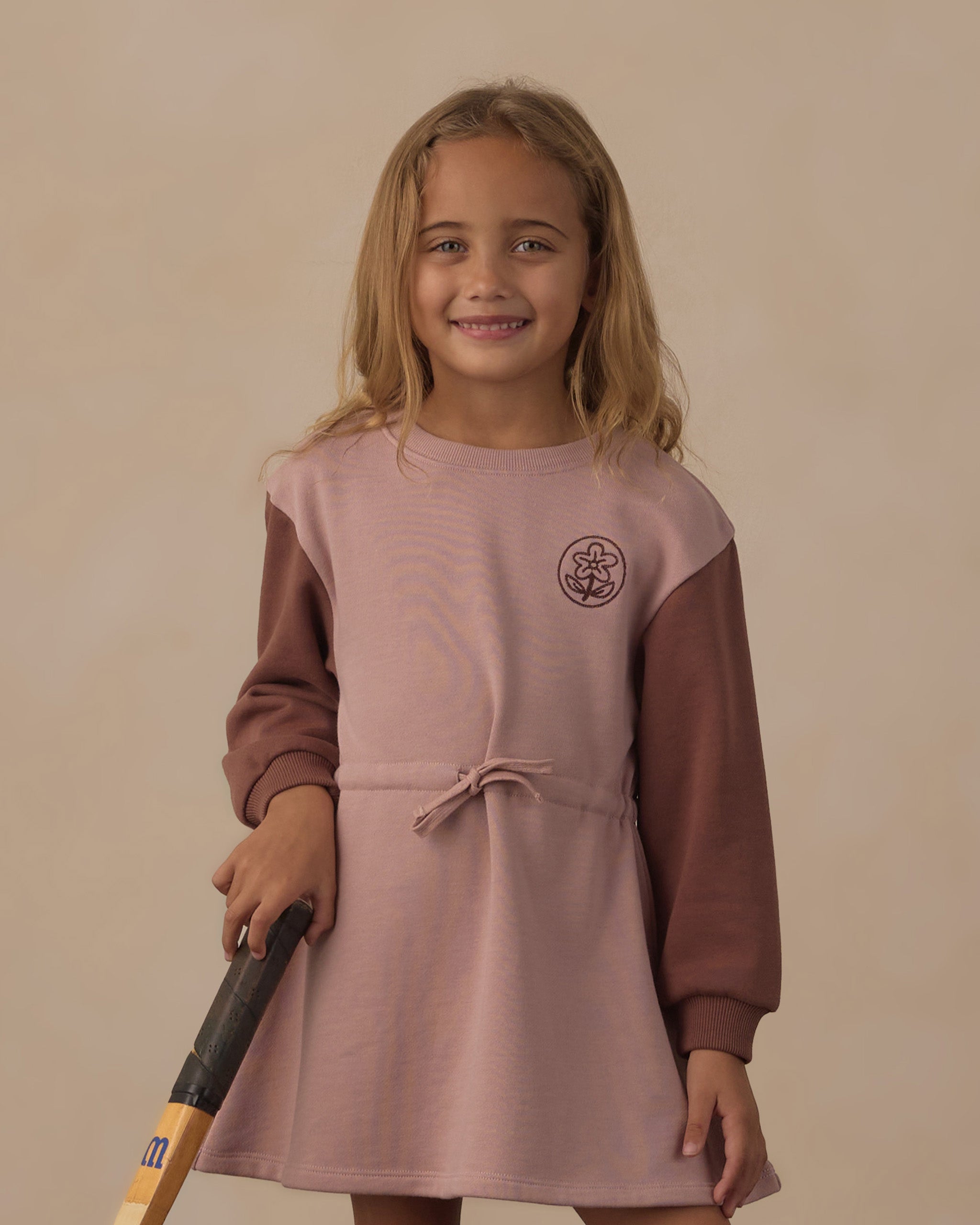 A young girl with blonde hair smiling and holding a baseball bat, wearing a long-sleeve dress with a dusty pink body and brown sleeves, set against a plain beige background.
