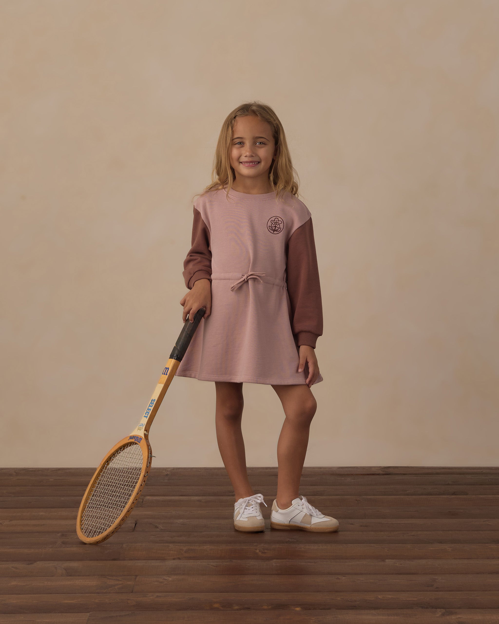 A young girl stands on a wooden floor against a beige wall, holding a tennis racket. She is wearing a two-tone long-sleeve dress in dusty pink and brown, with white sneakers.