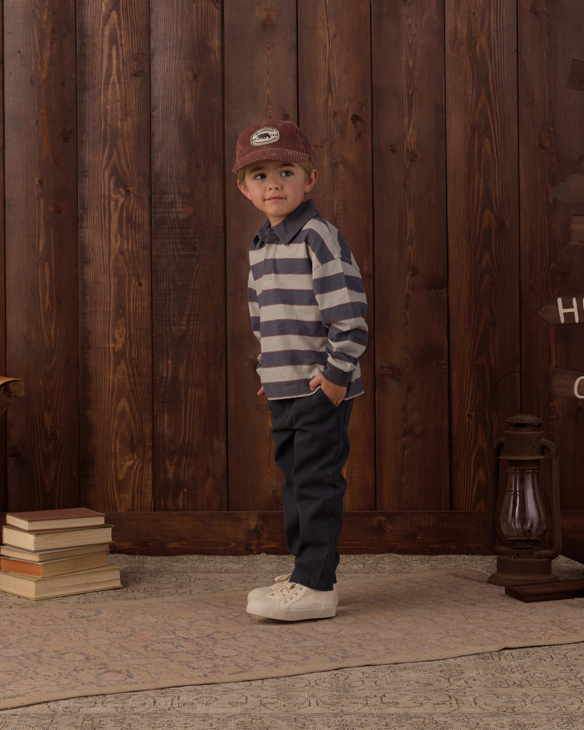A young boy stands in a rustic room with dark wood paneling, wearing a navy and gray striped long-sleeve shirt, dark pants, white sneakers, and a brown cap.