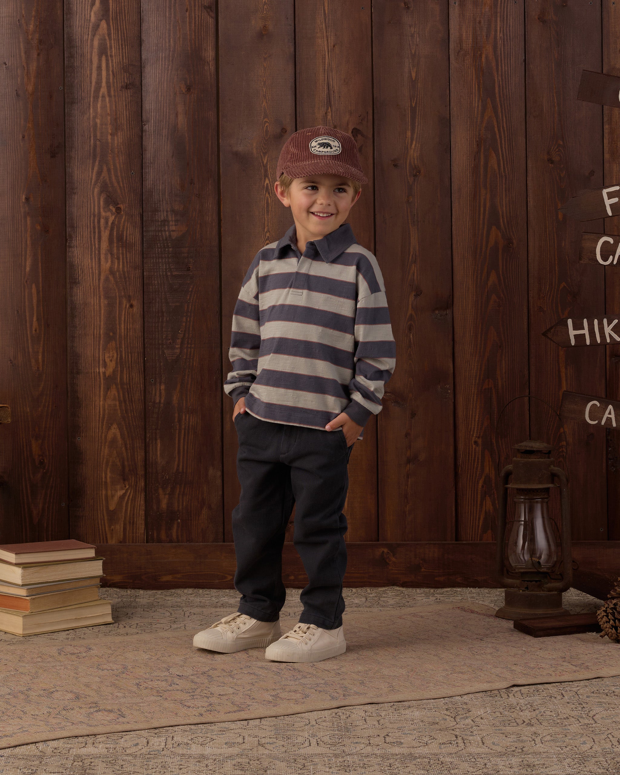 A young boy stands in front of a rustic dark wood plank wall wearing a navy and beige horizontally striped long-sleeve polo shirt, dark navy pants, off-white sneakers, and a brown corduroy cap. The setting includes a stone floor, a stack of books, a lantern, and wooden directional signs.