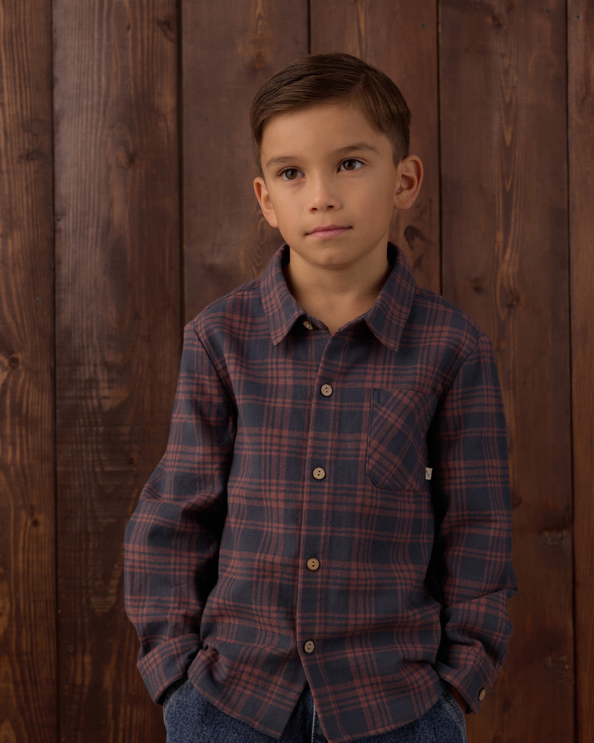 A young boy wearing a long-sleeve button-down plaid shirt in dark blue and red, standing against a dark brown wooden plank wall.