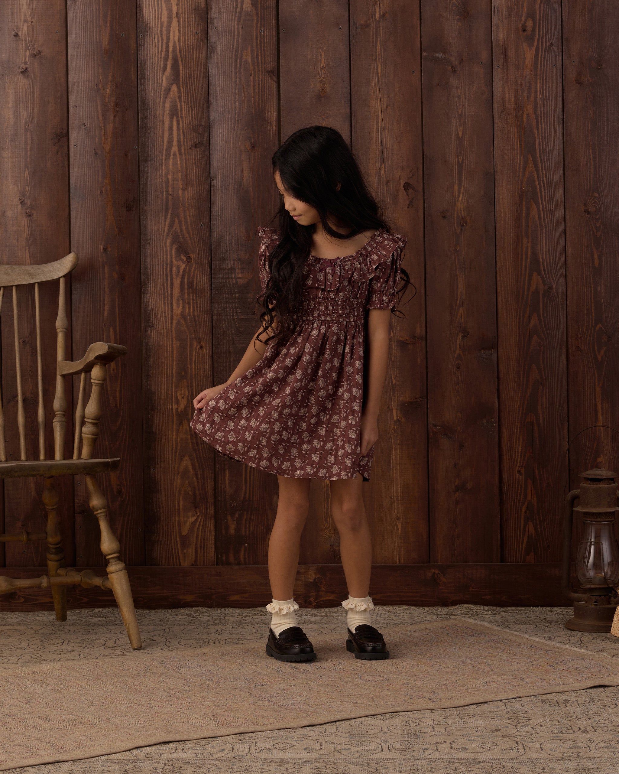 A young girl with long dark hair stands in front of a rustic dark wood plank wall, wearing a short-sleeved dusty rose and white floral print dress, white socks, and black shoes.