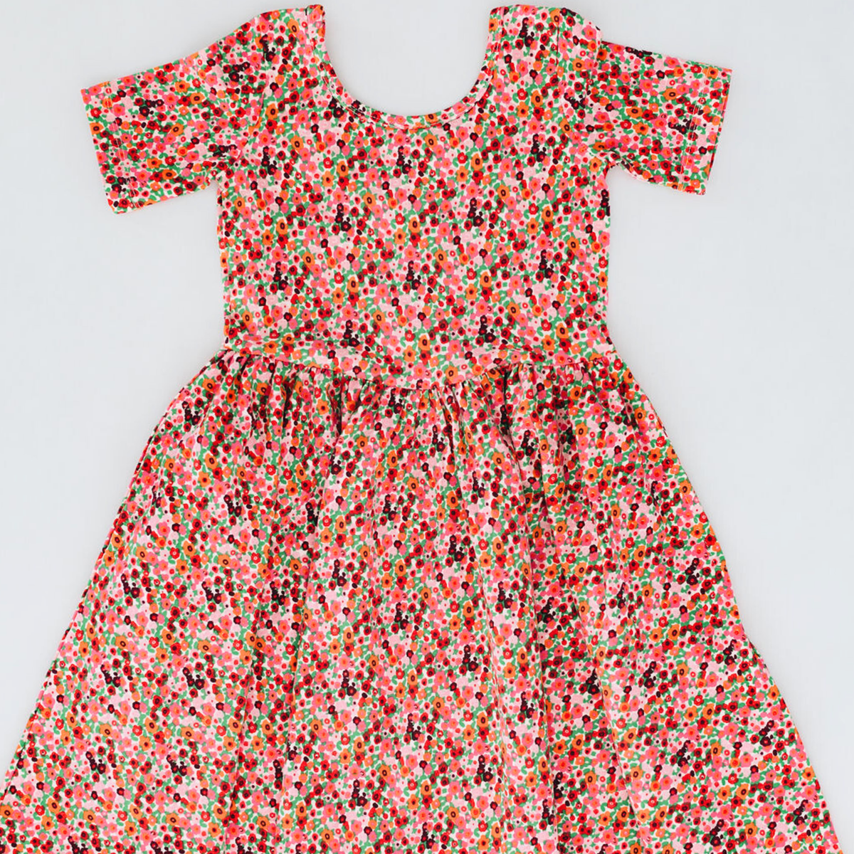A short-sleeve babydoll dress with a dense multicolor floral and abstract print on a light pink background, laid flat against a light gray background.