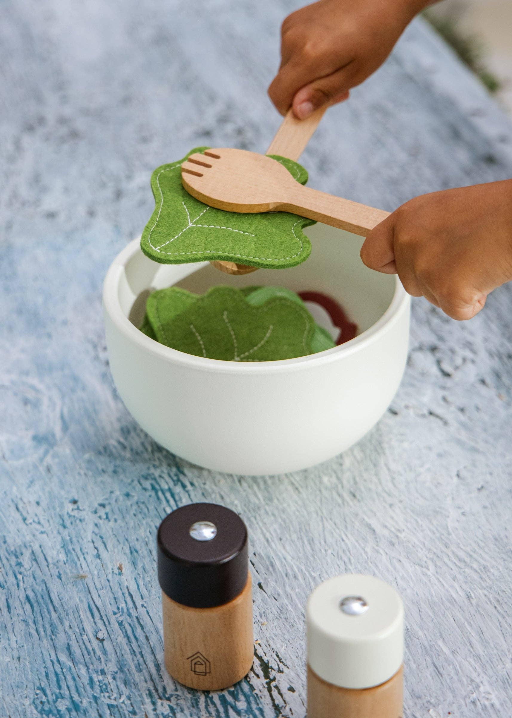 Children's play kitchen set with wooden spoon and leaf-shaped spatula over a white bowl.
