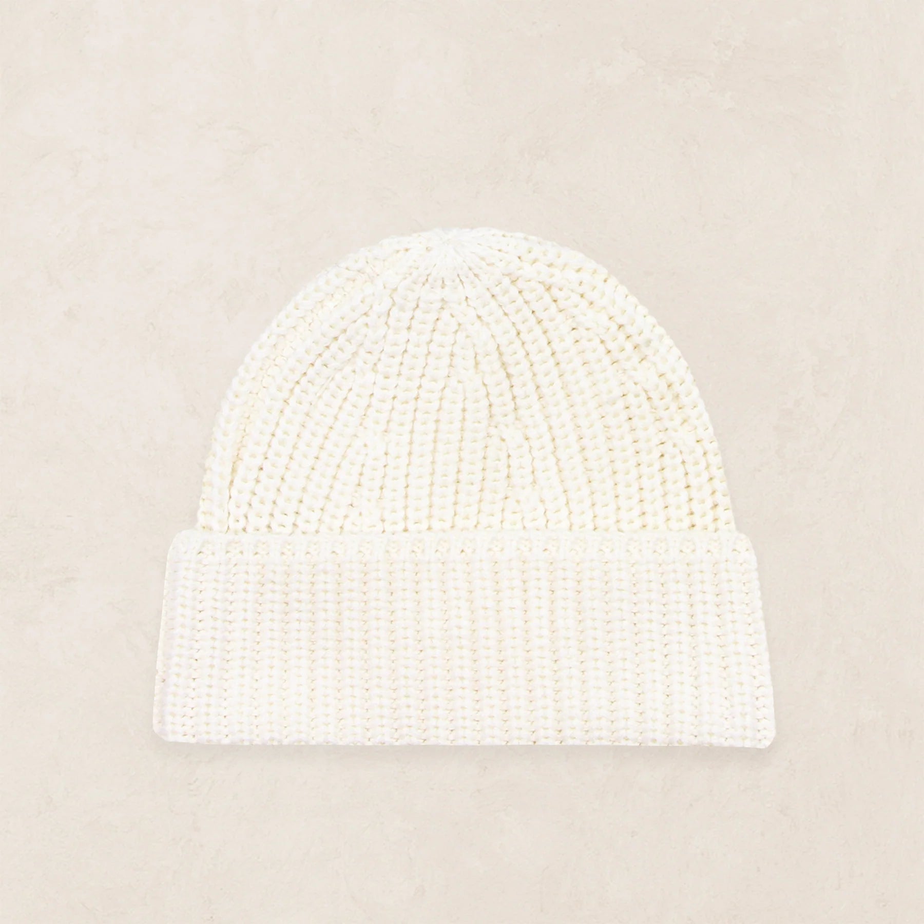 A cream-colored, ribbed knit beanie hat with a folded cuff, displayed flat against a textured beige background.