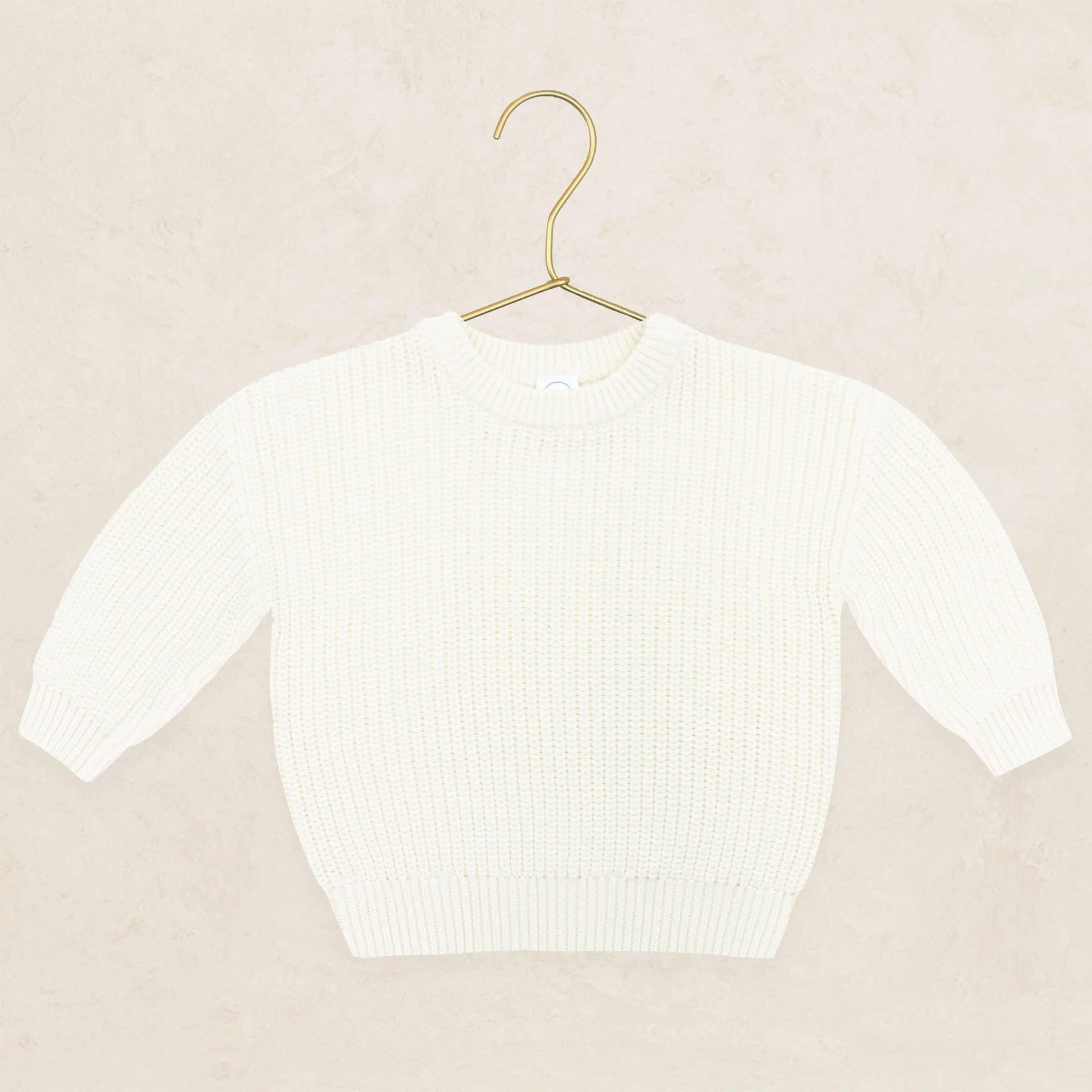 A cream-colored ribbed knit sweater displayed on a gold metal hanger against a textured beige background.